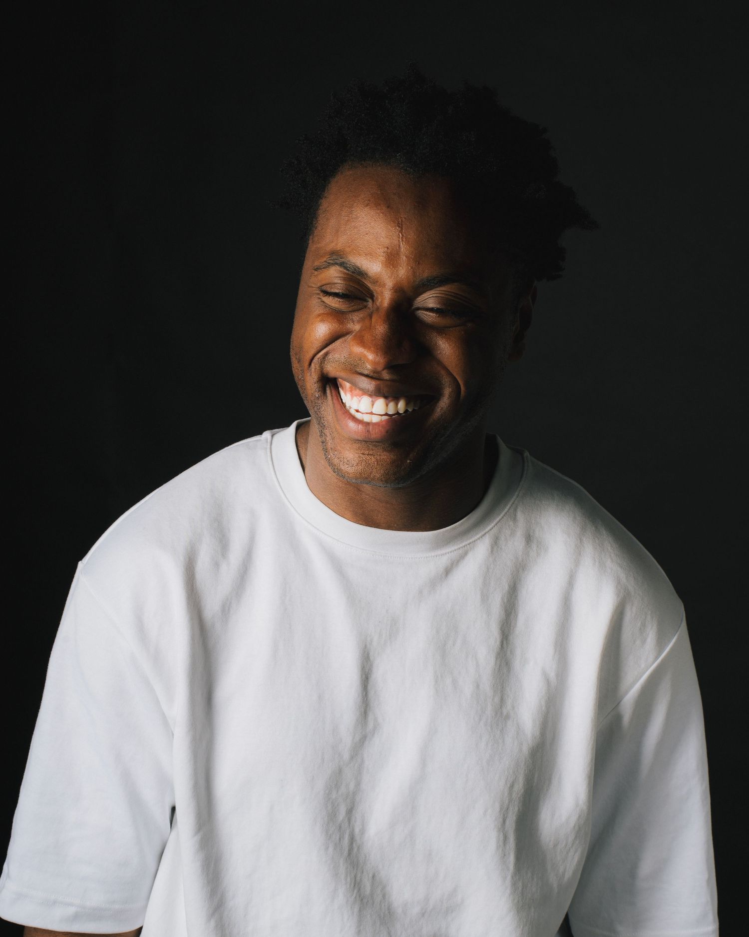 Joyful person wearing a white t-shirt laughs genuinely against a dark background in a portrait style photo.