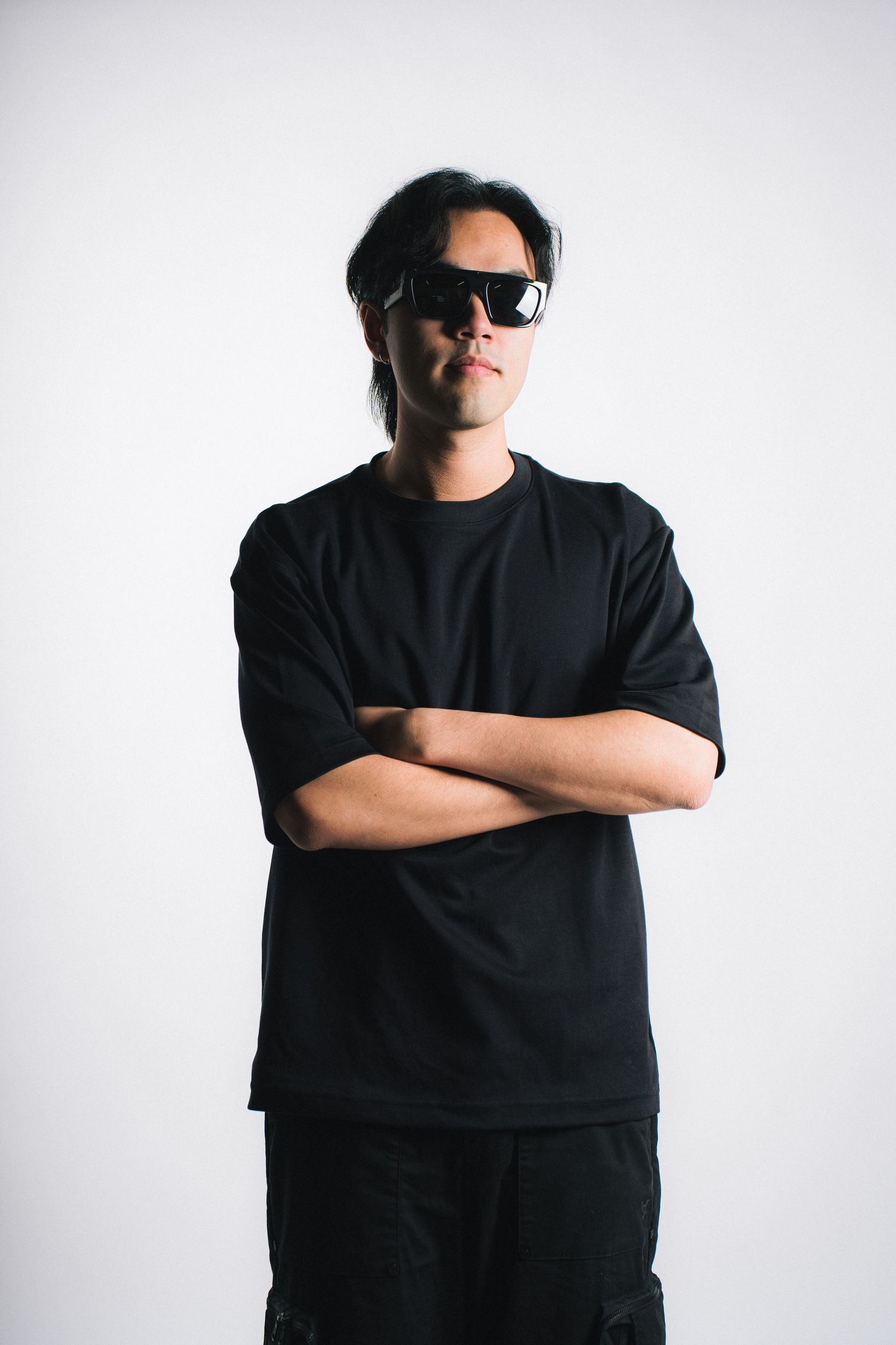 A person wearing a black t-shirt and sunglasses poses against a white backdrop with arms crossed.