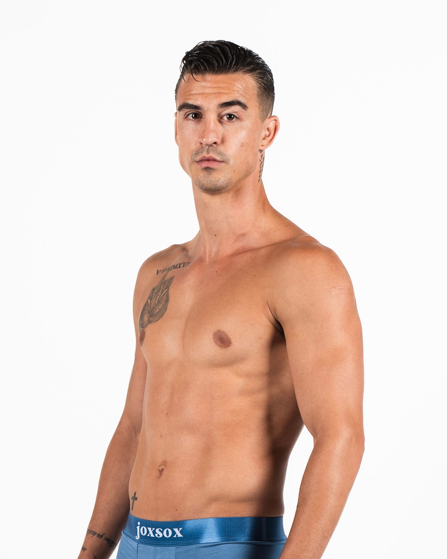 Athletic man with short dark hair poses shirtless wearing blue boxer briefs, showing tattoo on shoulder.