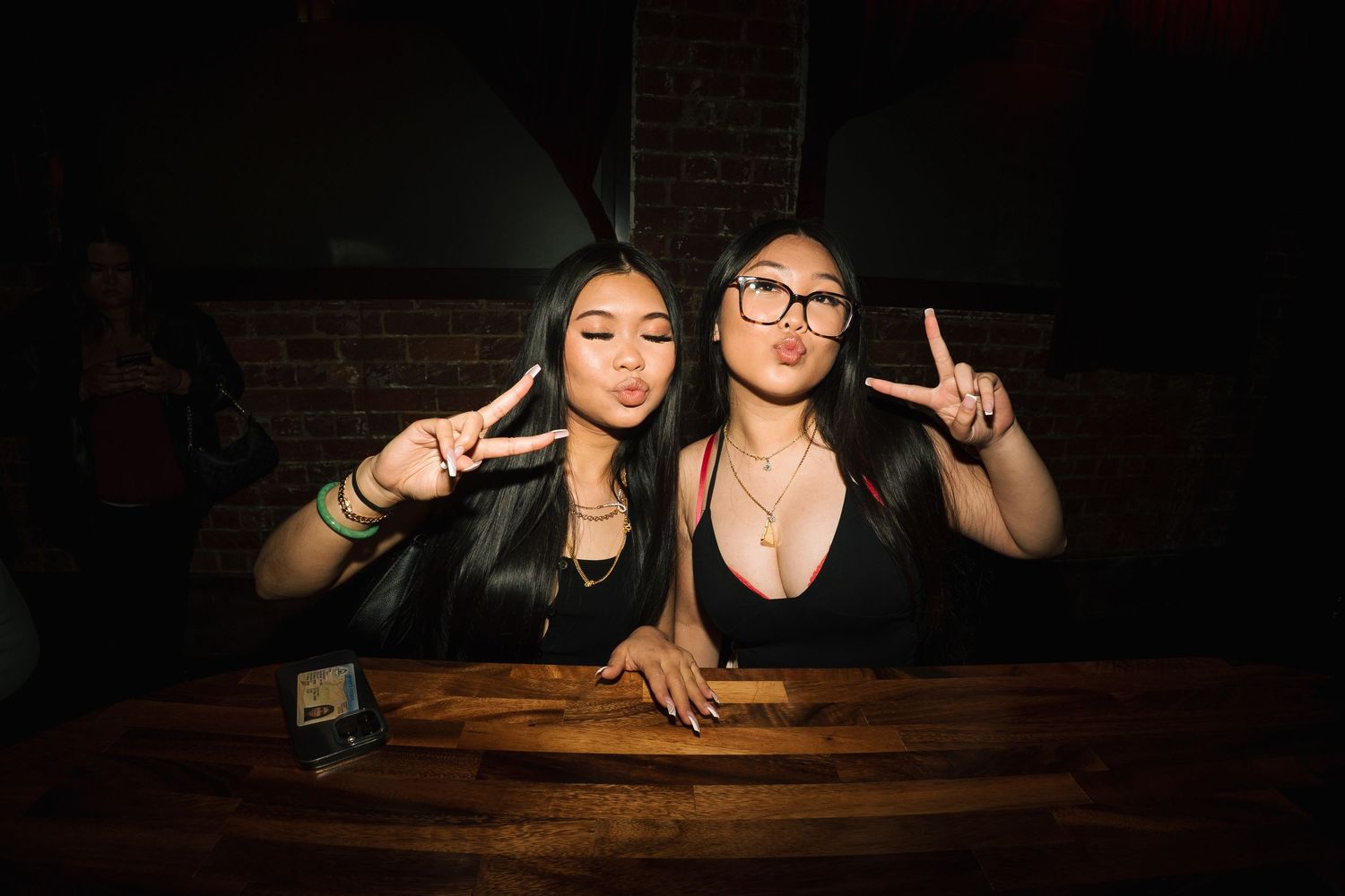 Two friends make peace signs while sitting at a dark wooden table in a dimly lit room.