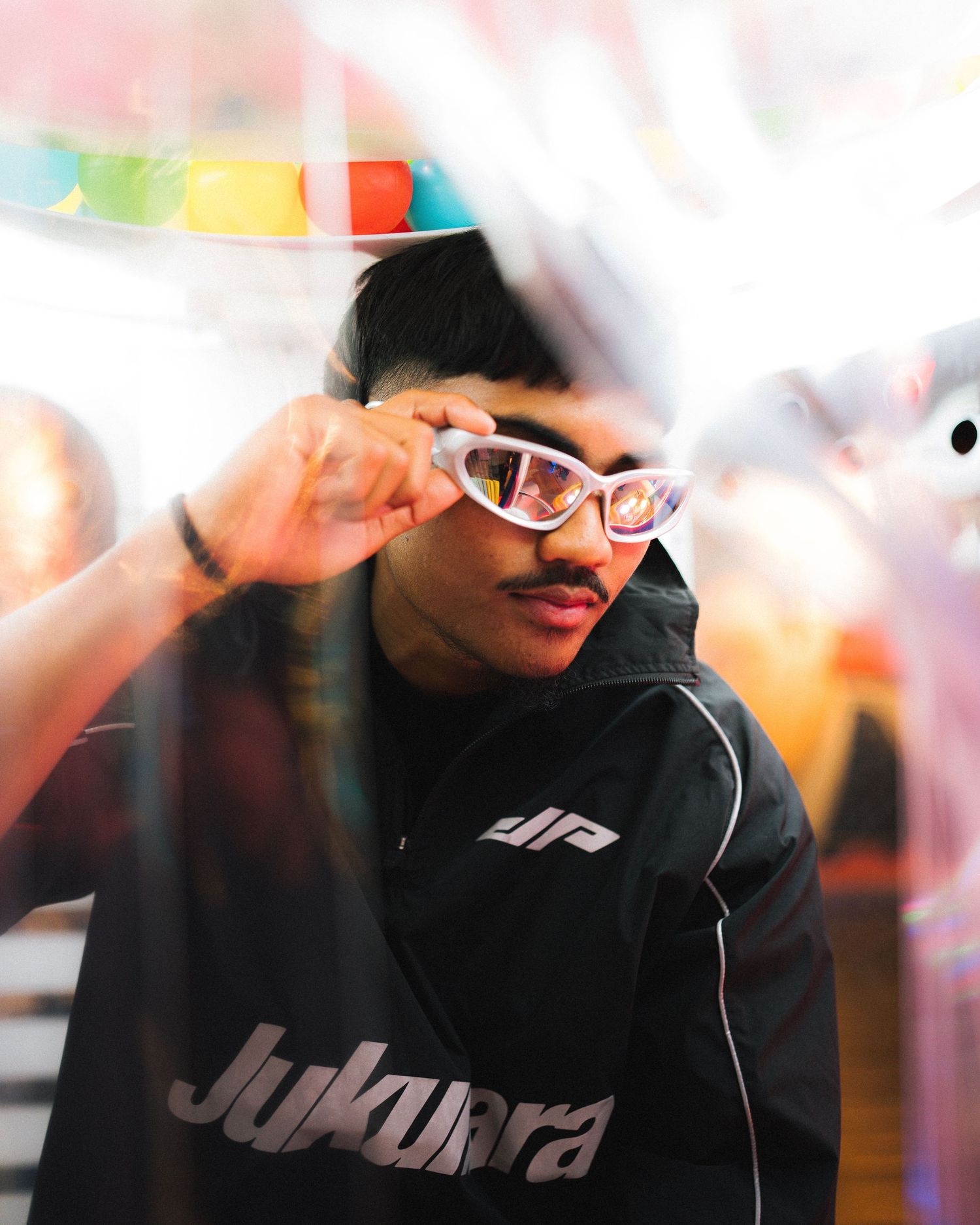 A person in a black hoodie adjusts their white-rimmed glasses in a soft-focus party setting with colorful decorations.
