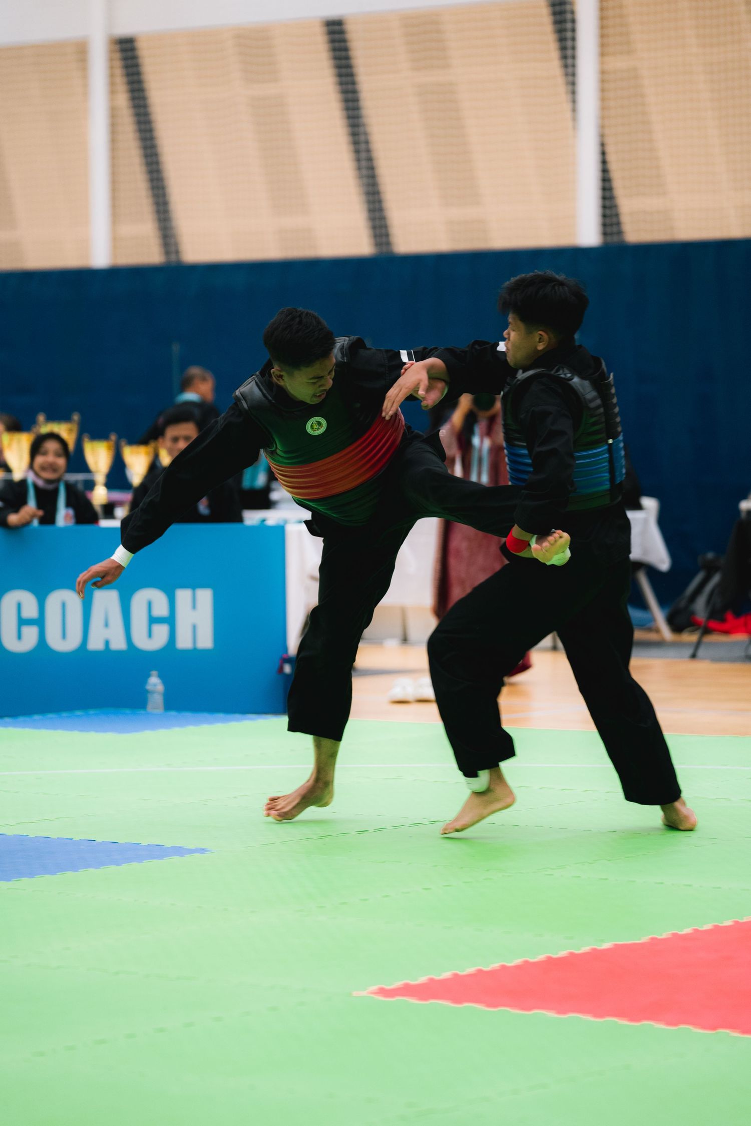 Two martial artists in black uniforms practicing kicks during a sparring match on colorful mats.