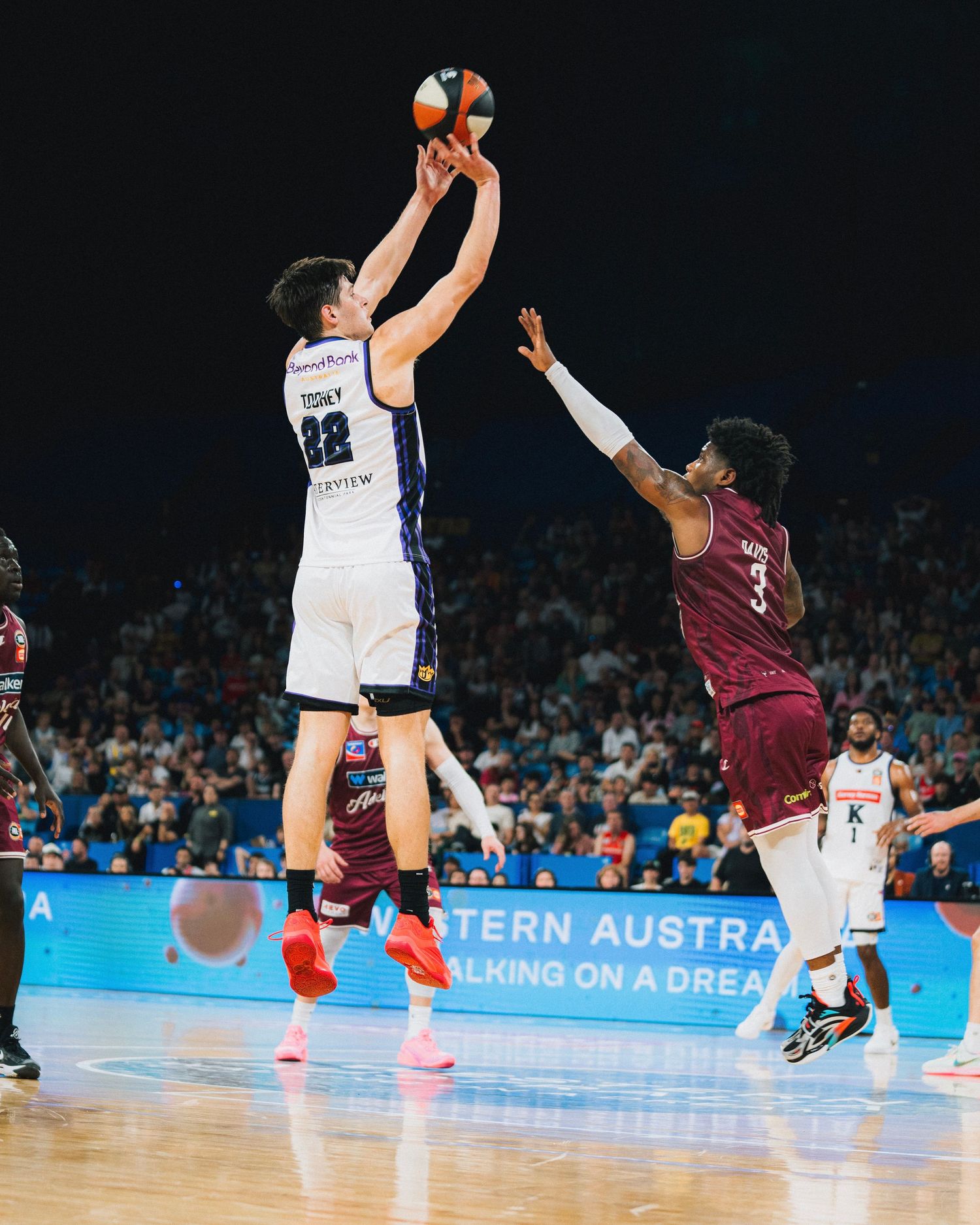 Basketball player takes a jump shot during a competitive game as defender tries to block.