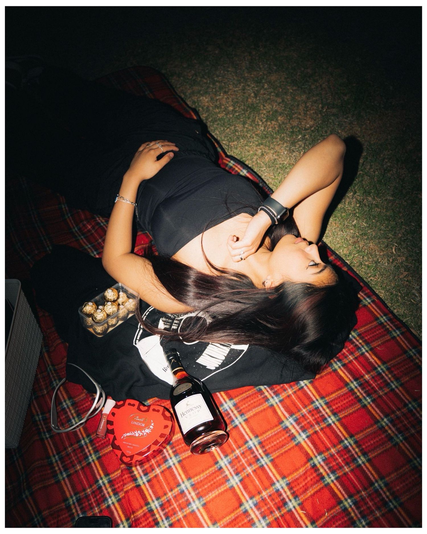 Person lying on red plaid blanket with camera and accessories at night.