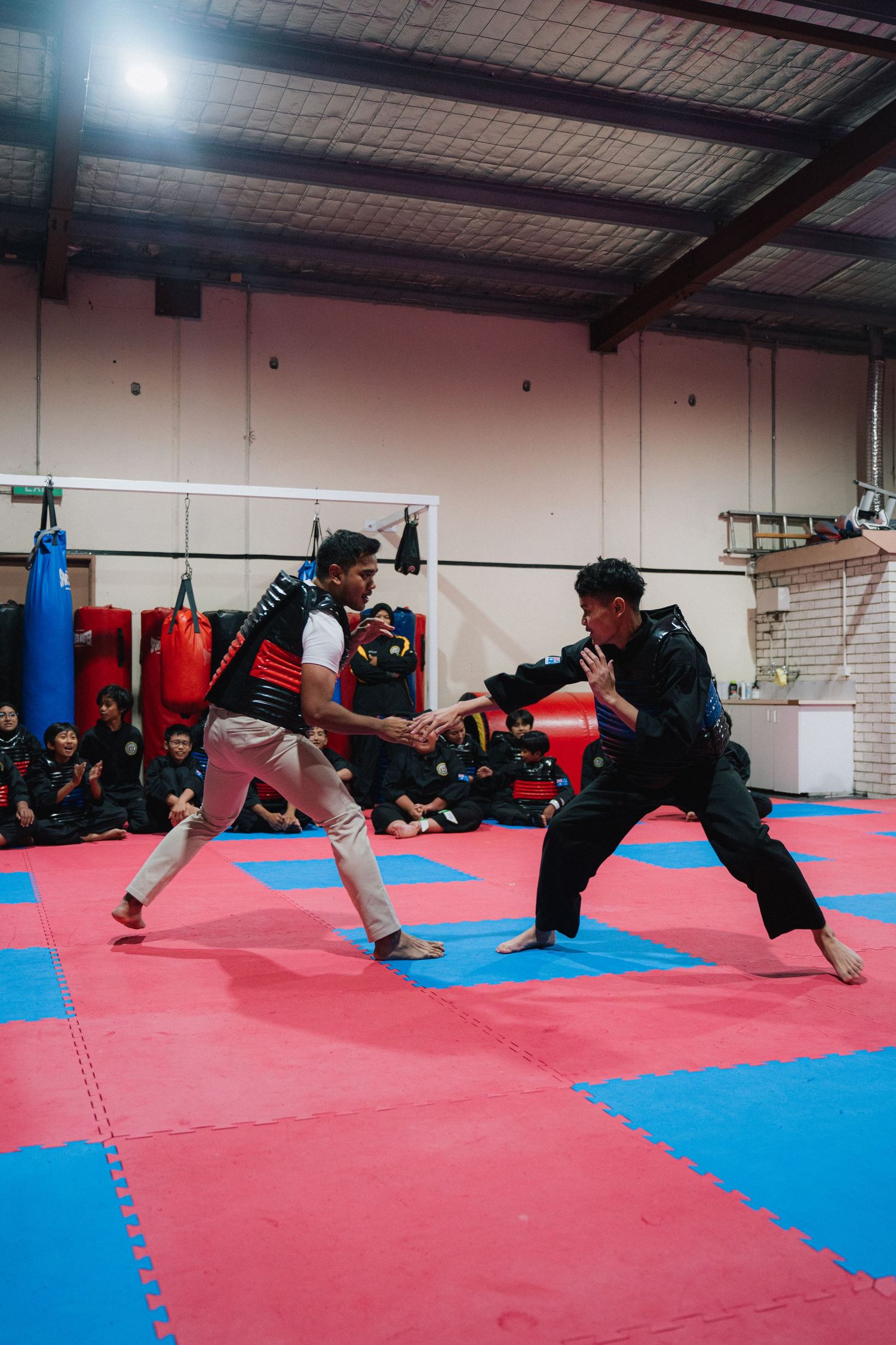 Two martial artists practicing sparring techniques on red and blue mats in a training gym.