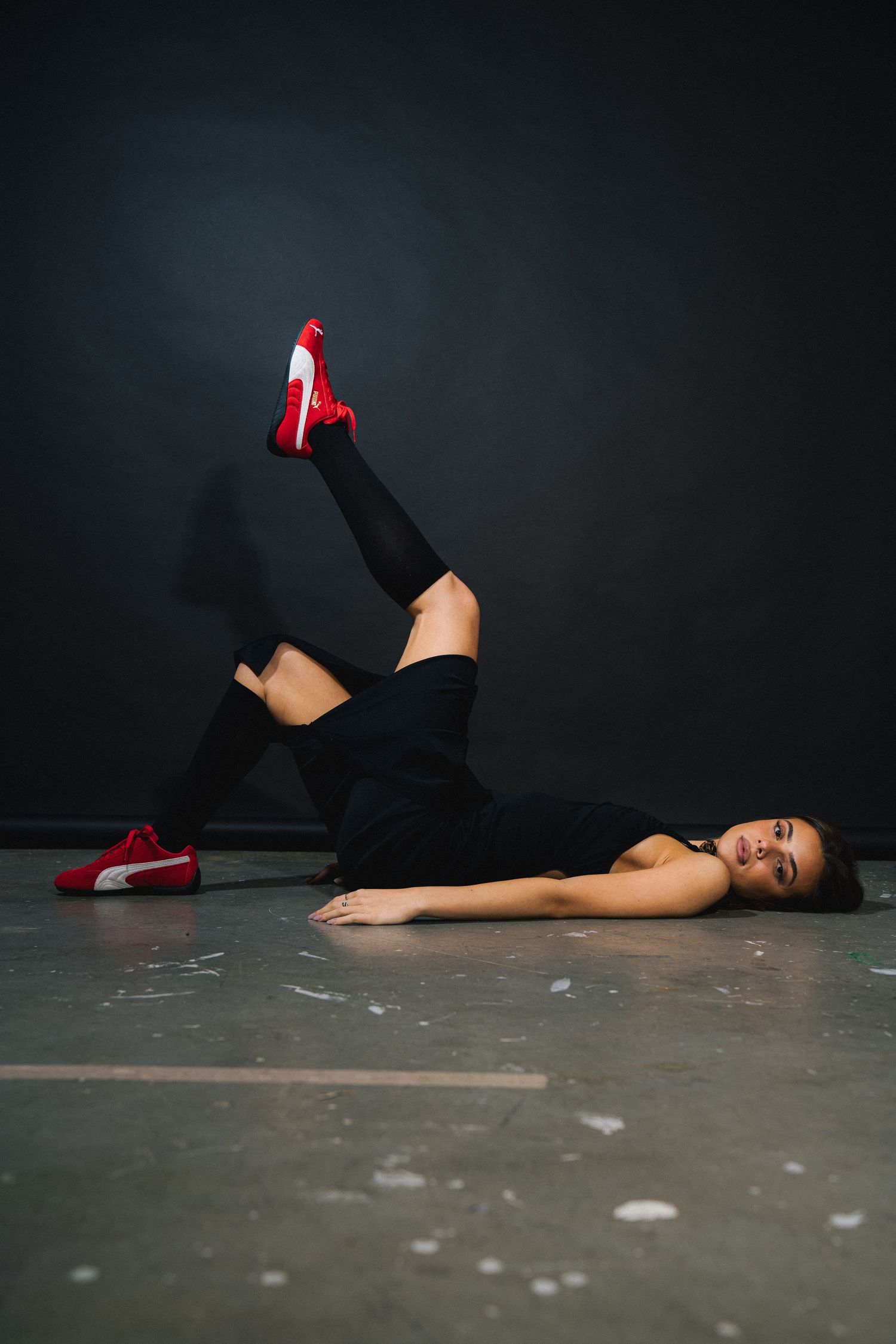 Dancer in black outfit with red shoes performing floor routine against dark background.