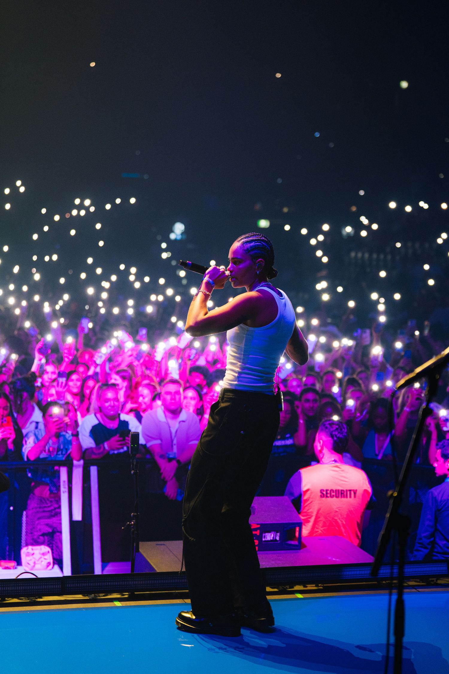 Concert performer silhouetted on stage with purple lights and phone lights glowing in large crowd background.