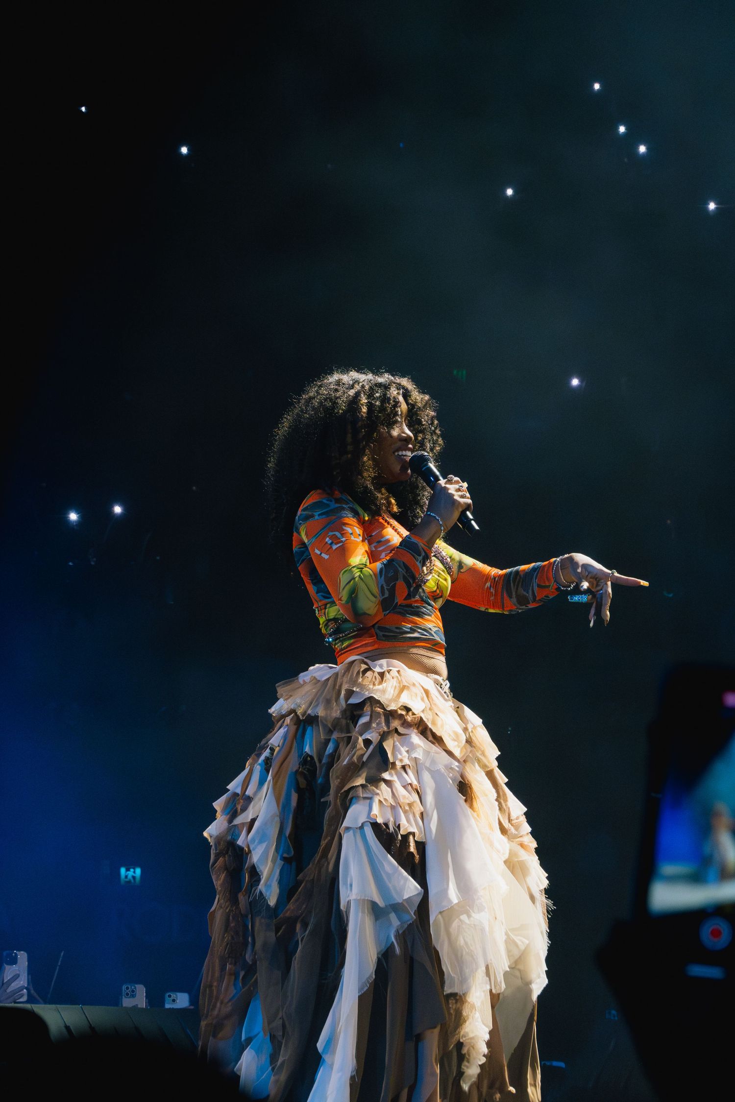 A performer in a colorful top and layered white dress sings on stage with dramatic lighting and sparkles in the background.