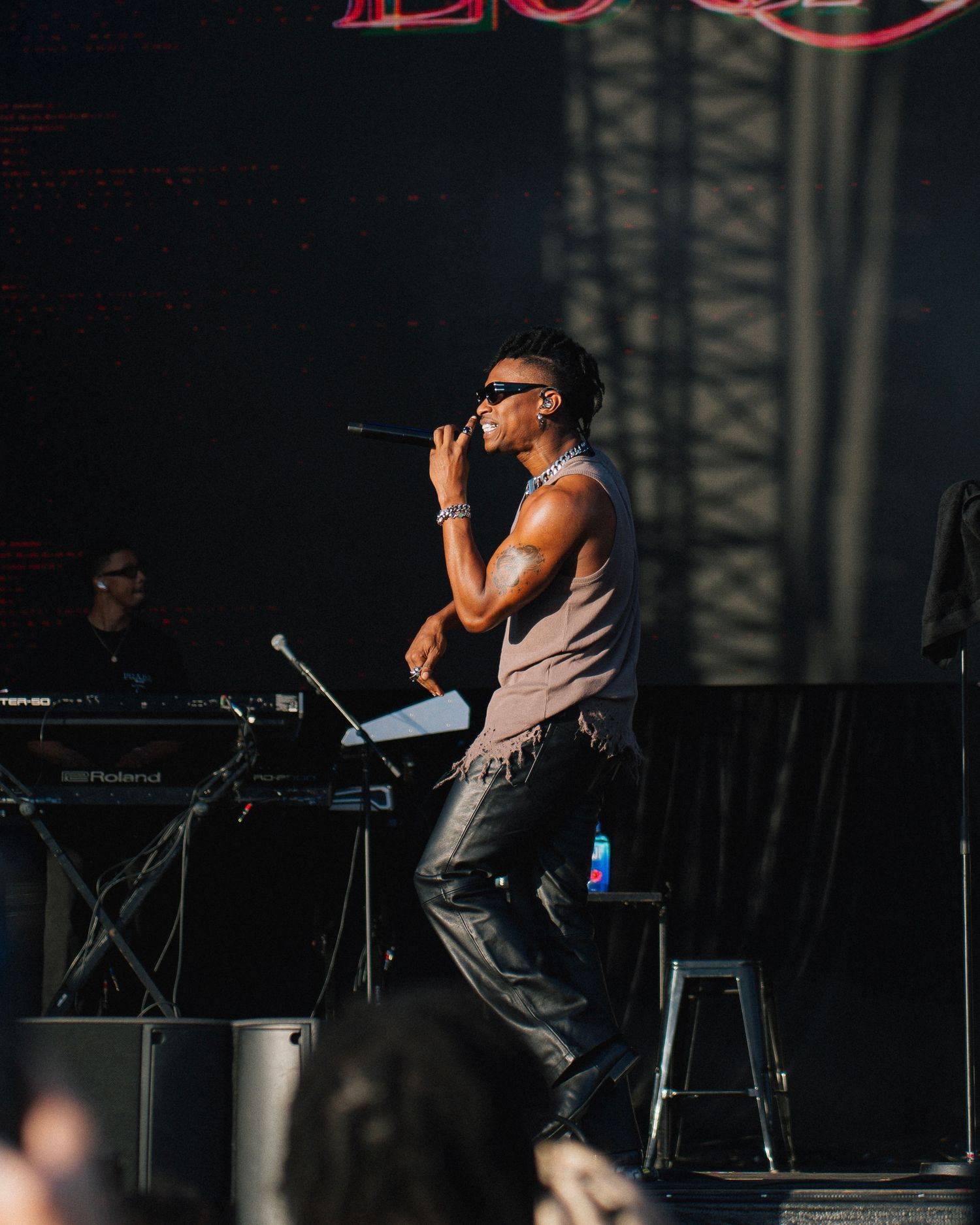 A performer in a tank top and sunglasses sings passionately on stage with a keyboard player visible in the background.