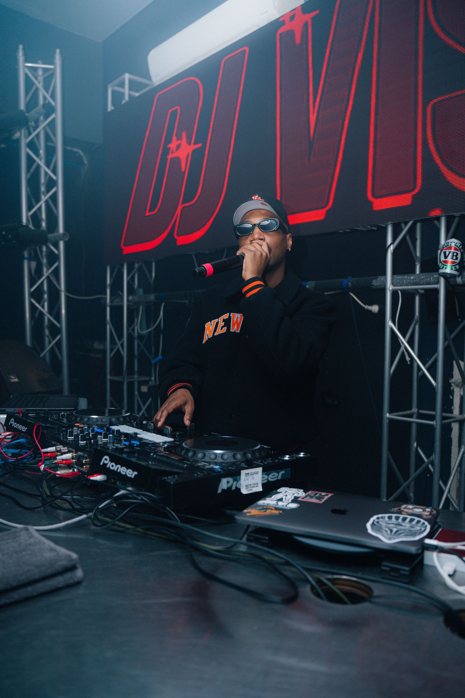DJ performing under red neon signage with Pioneer equipment in dark club setting.