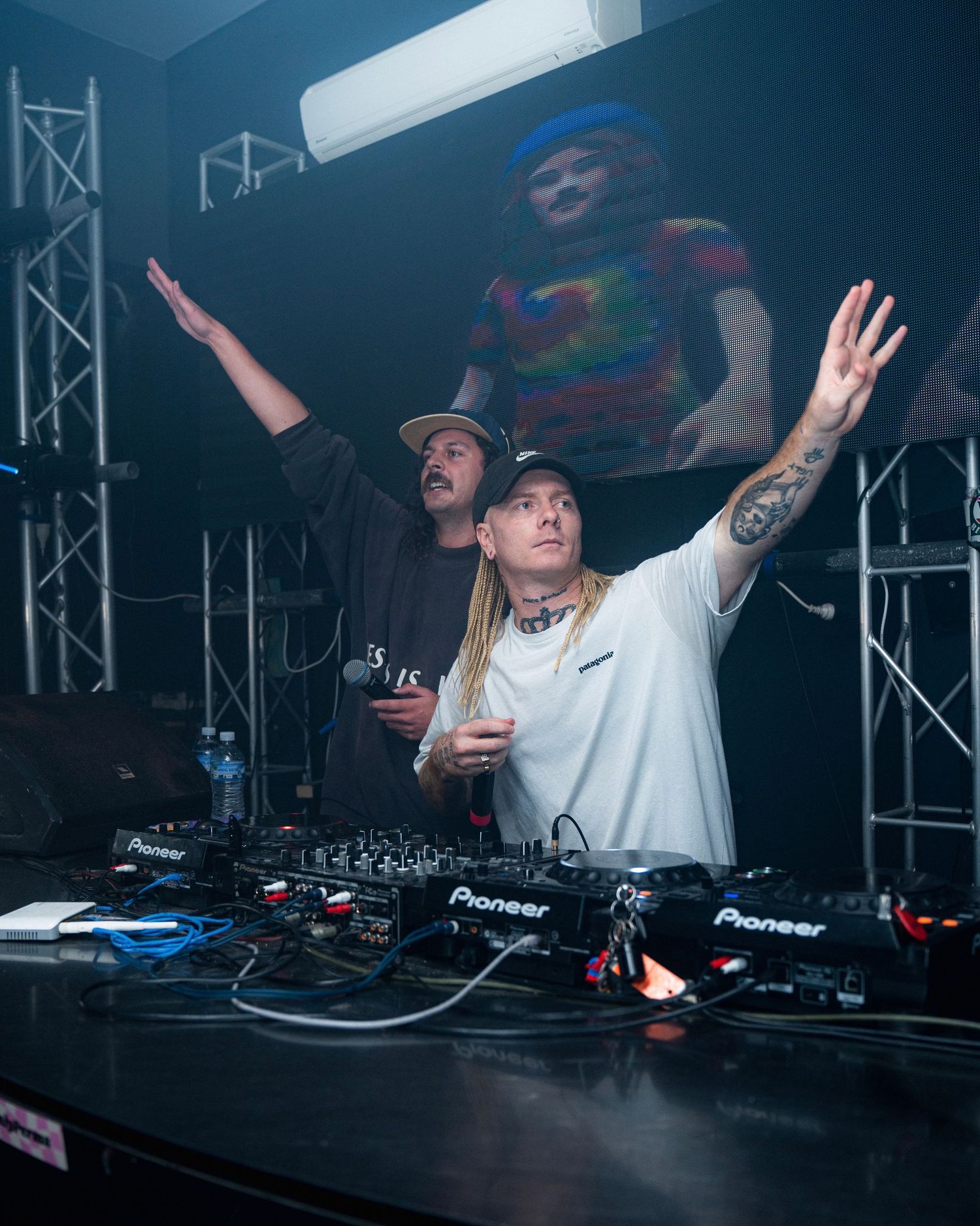 DJs performing enthusiastically behind Pioneer decks with stage lighting and raised hands.