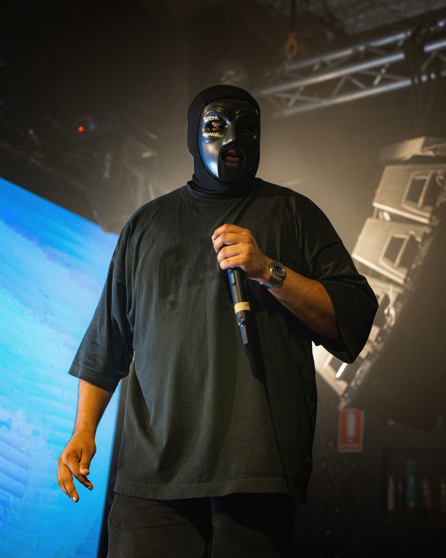 Masked performer in black clothing holding microphone on stage with dramatic lighting.