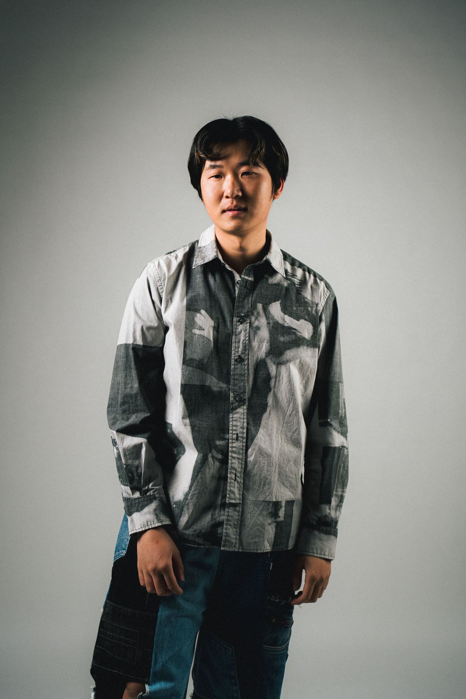 Portrait of a person wearing a casual patchwork denim shirt and jeans standing against a grey background.