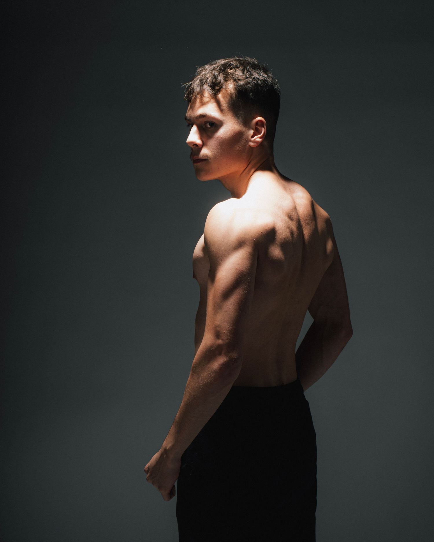 Dramatic studio portrait showing muscular shirtless figure in profile against dark background with dramatic lighting.