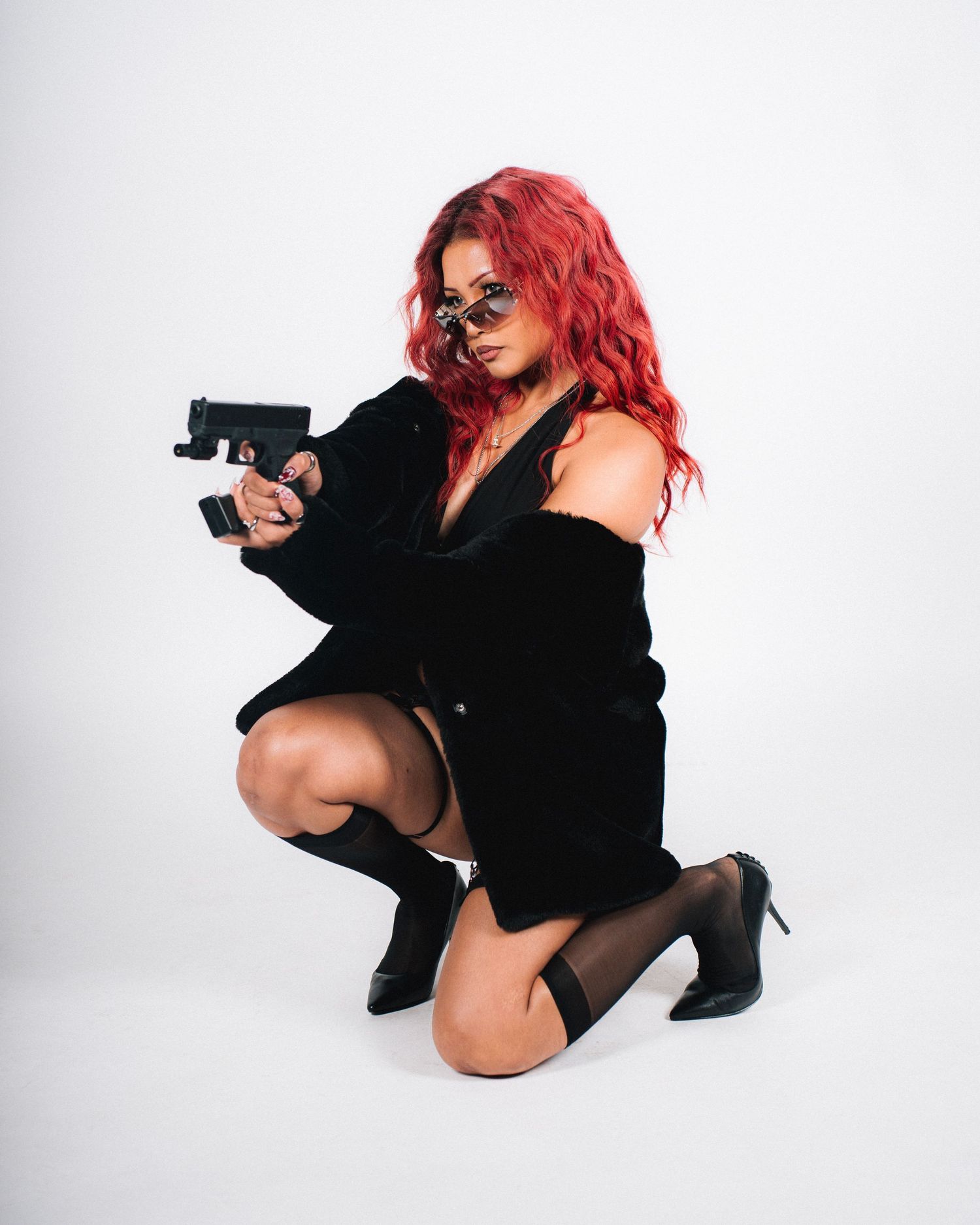 Fashion model in black outfit with bright hair poses in crouching position with prop against white backdrop while wearing sunglasses.