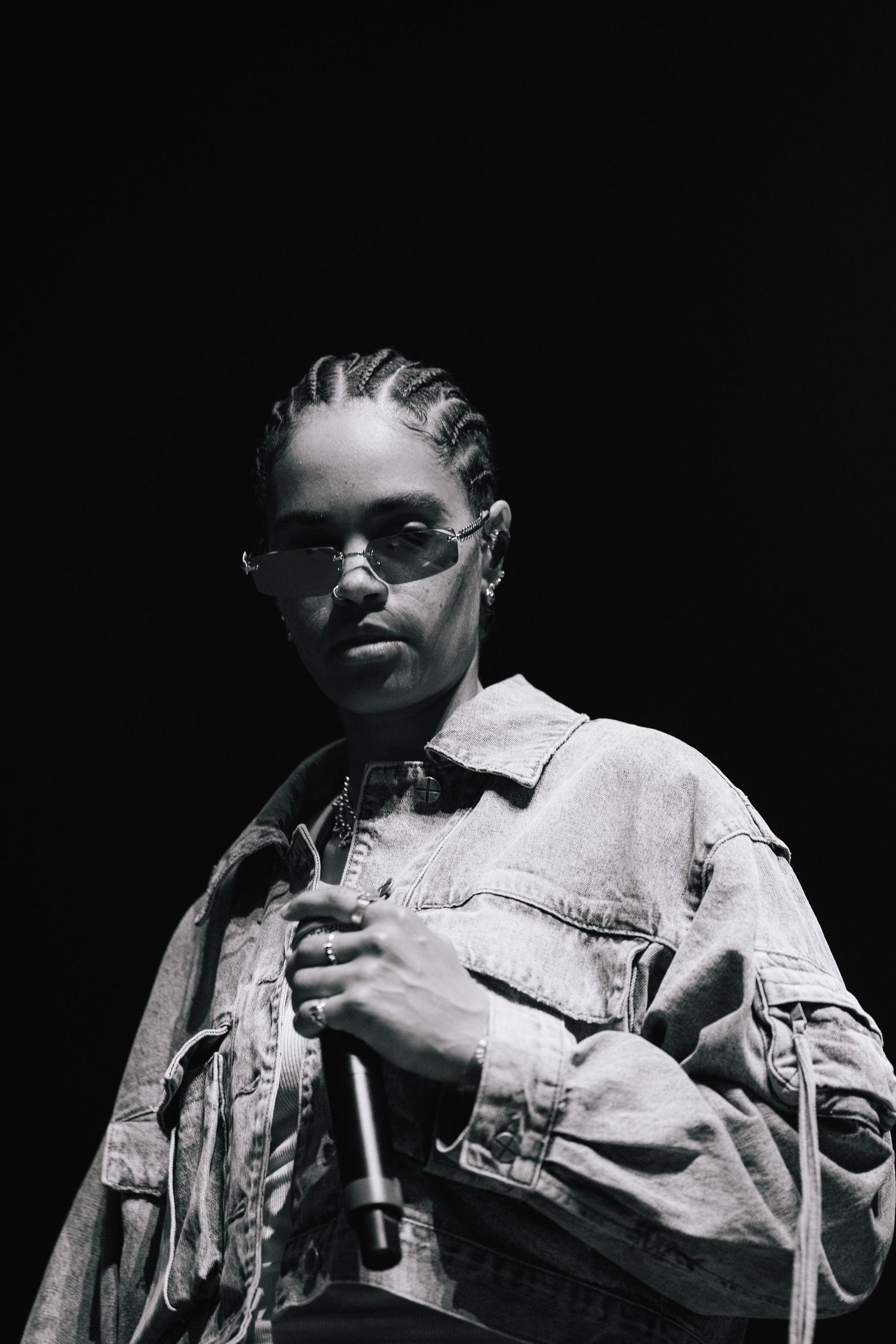 A performer in cornrow braids wearing sunglasses and a light denim jacket holds a microphone in a dark setting.