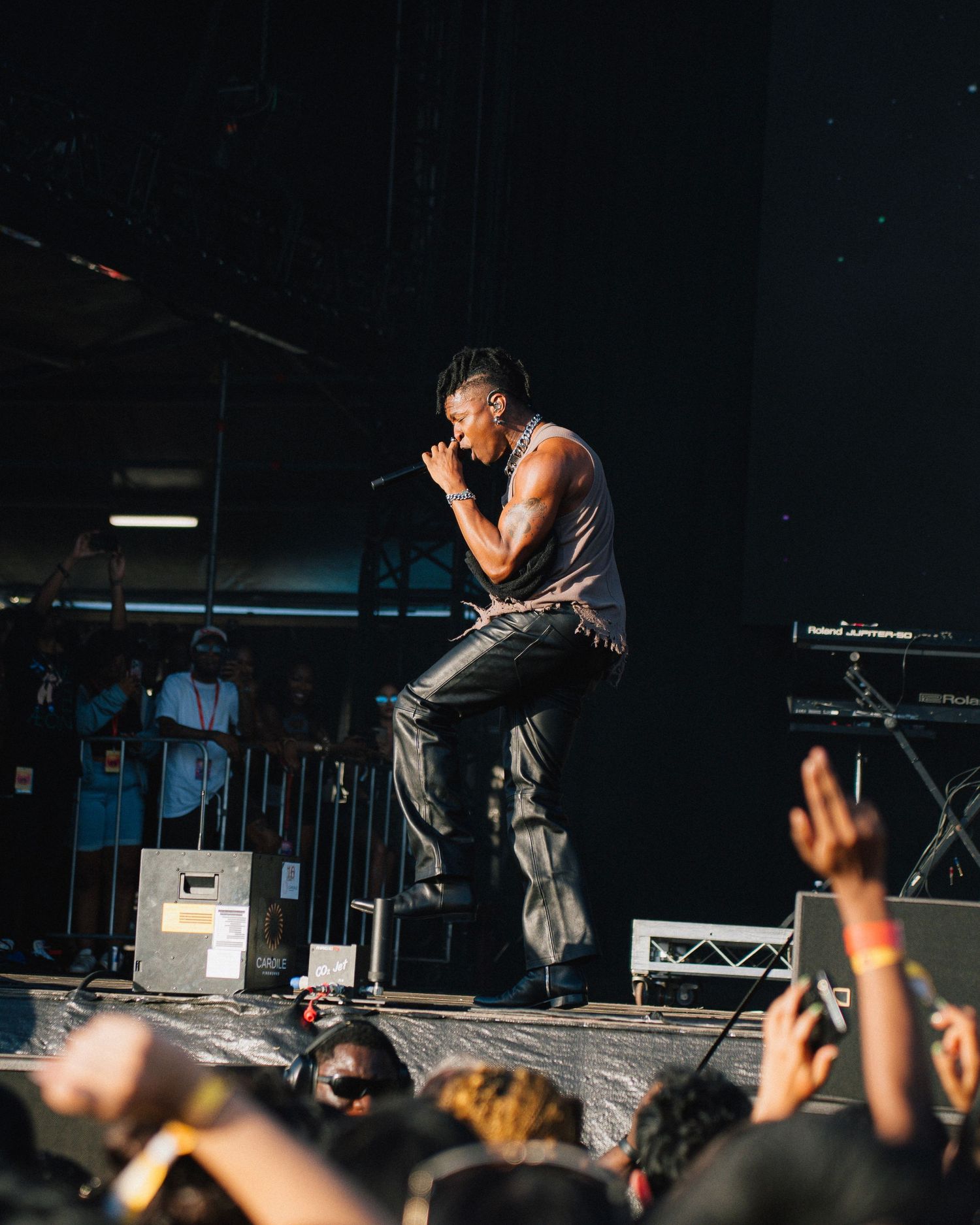 Performer on stage energetically singing into microphone at concert with crowd raising hands in dark venue during show.