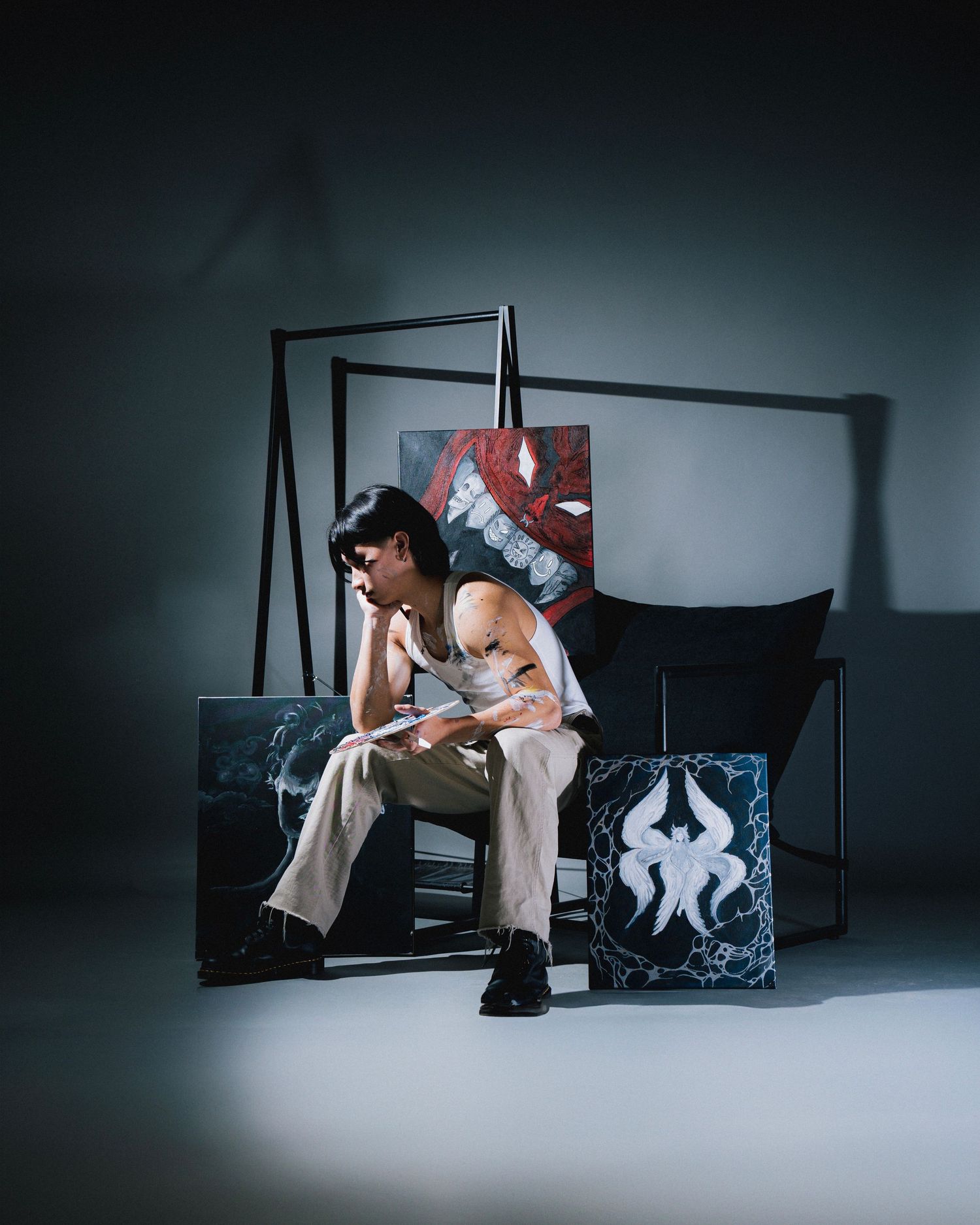 Artist sitting on bench with artwork in moody studio setting.