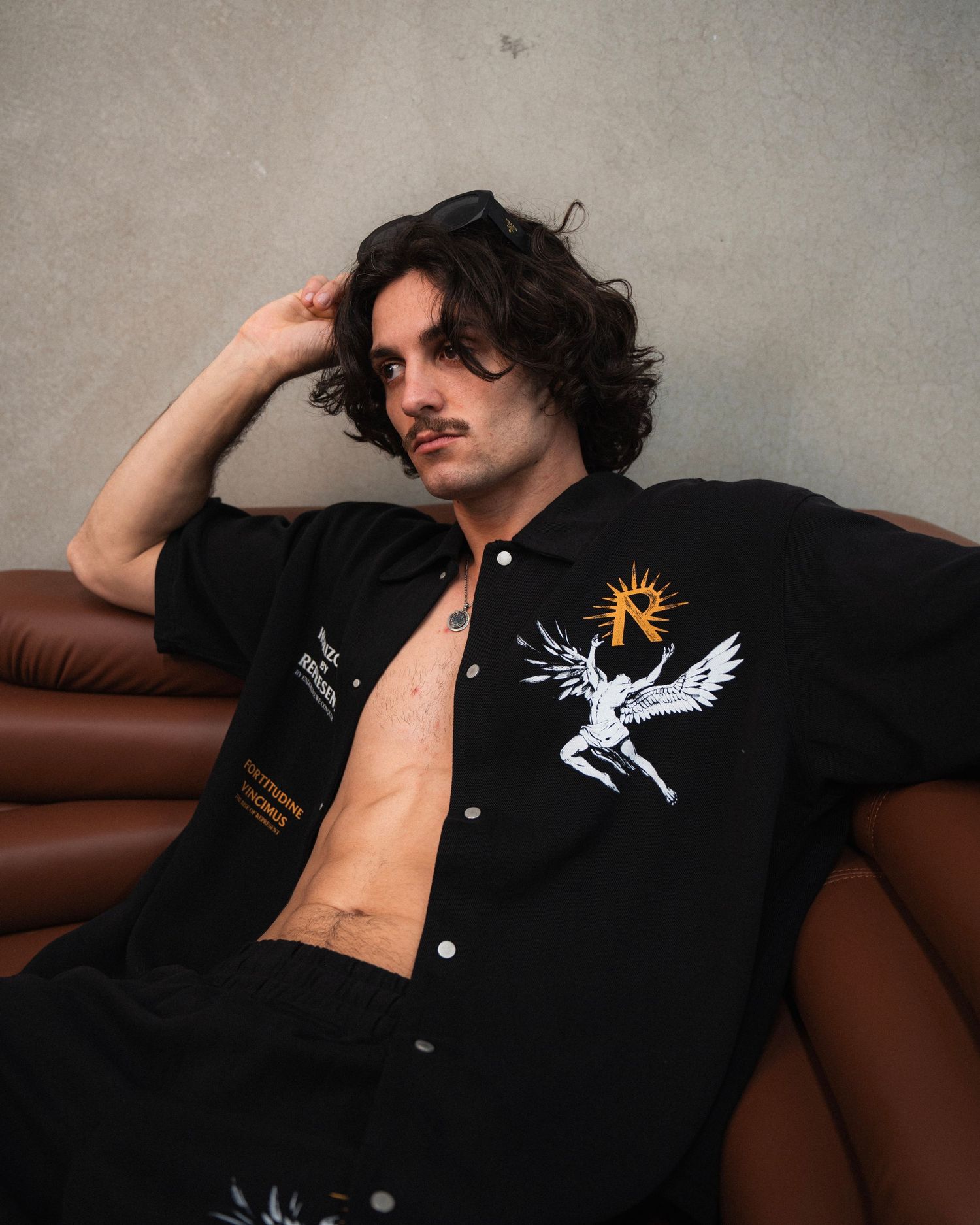 Model in open black shirt with angel design lounges on brown leather sofa.