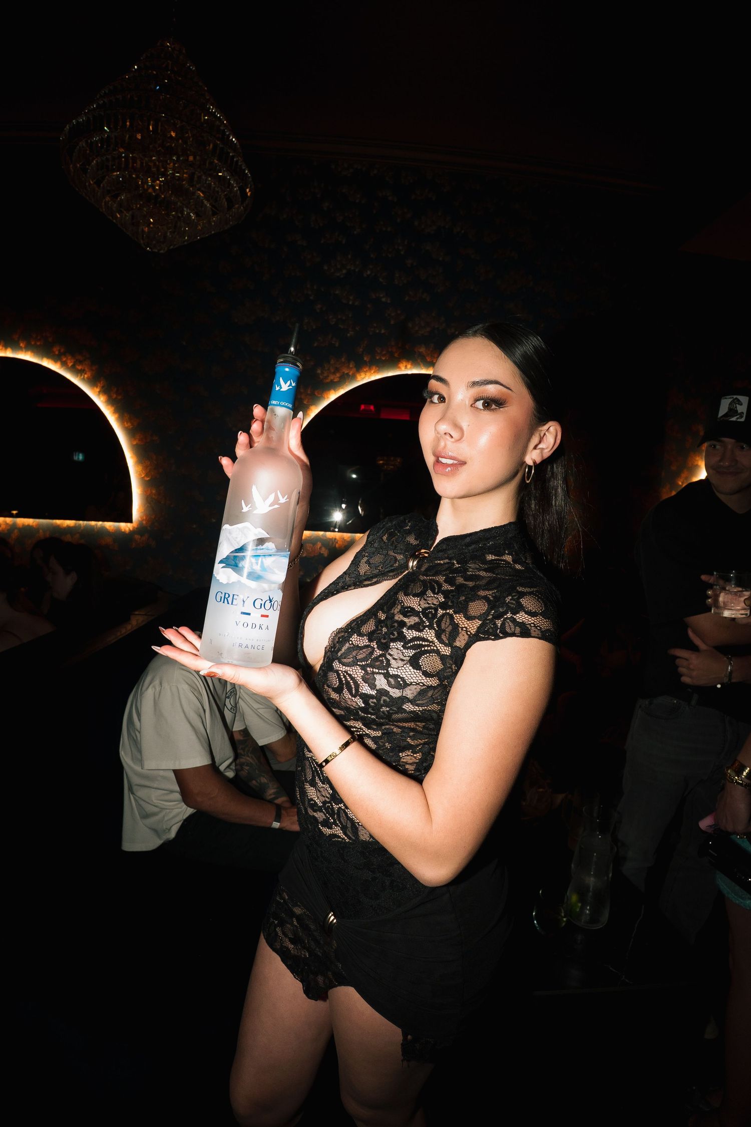 A woman in a black lace dress holds a bottle of Grey Goose vodka at a nightclub in dark lighting.