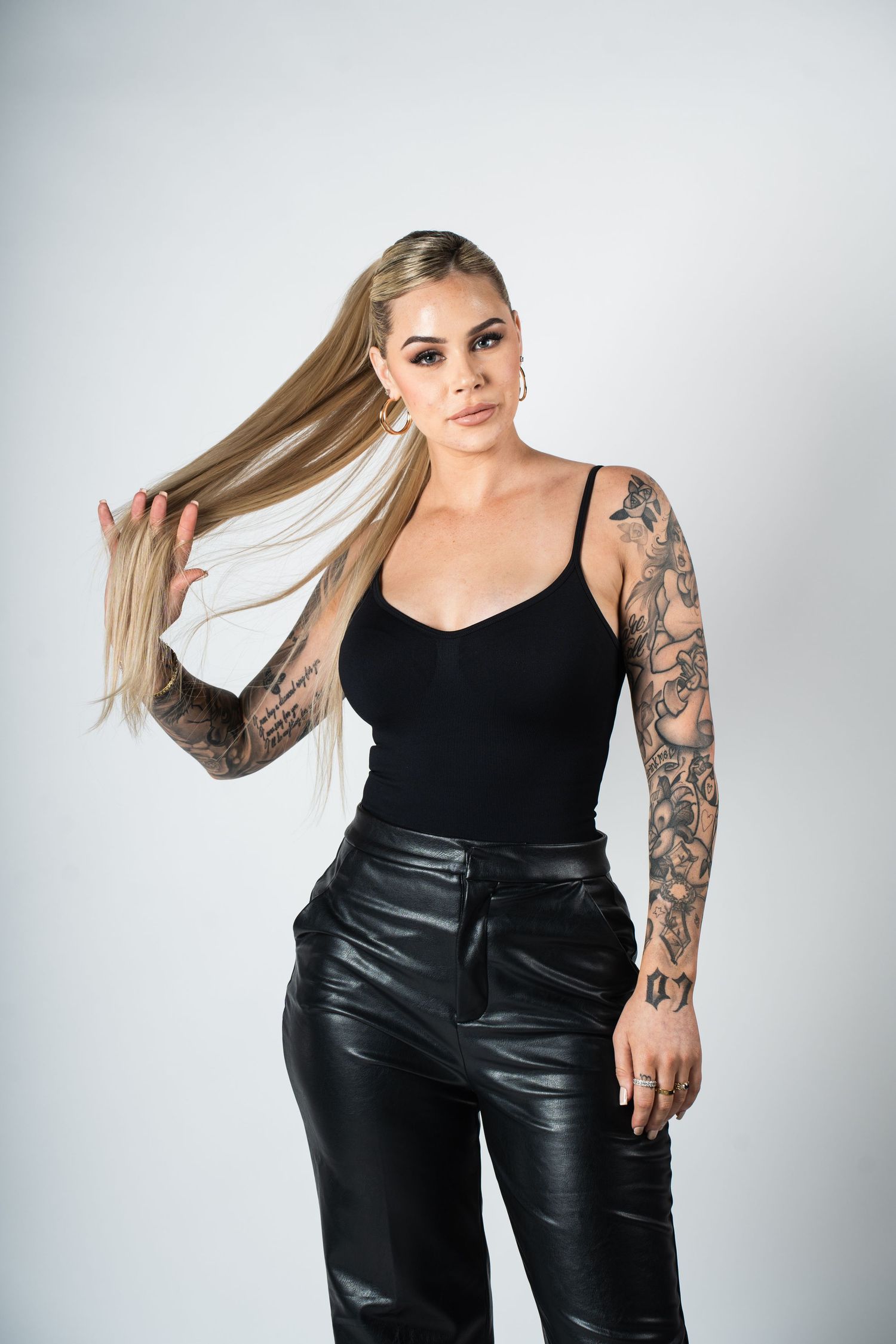 Stylish fashion model with tattoos poses in black tank top and leather pants against light background.