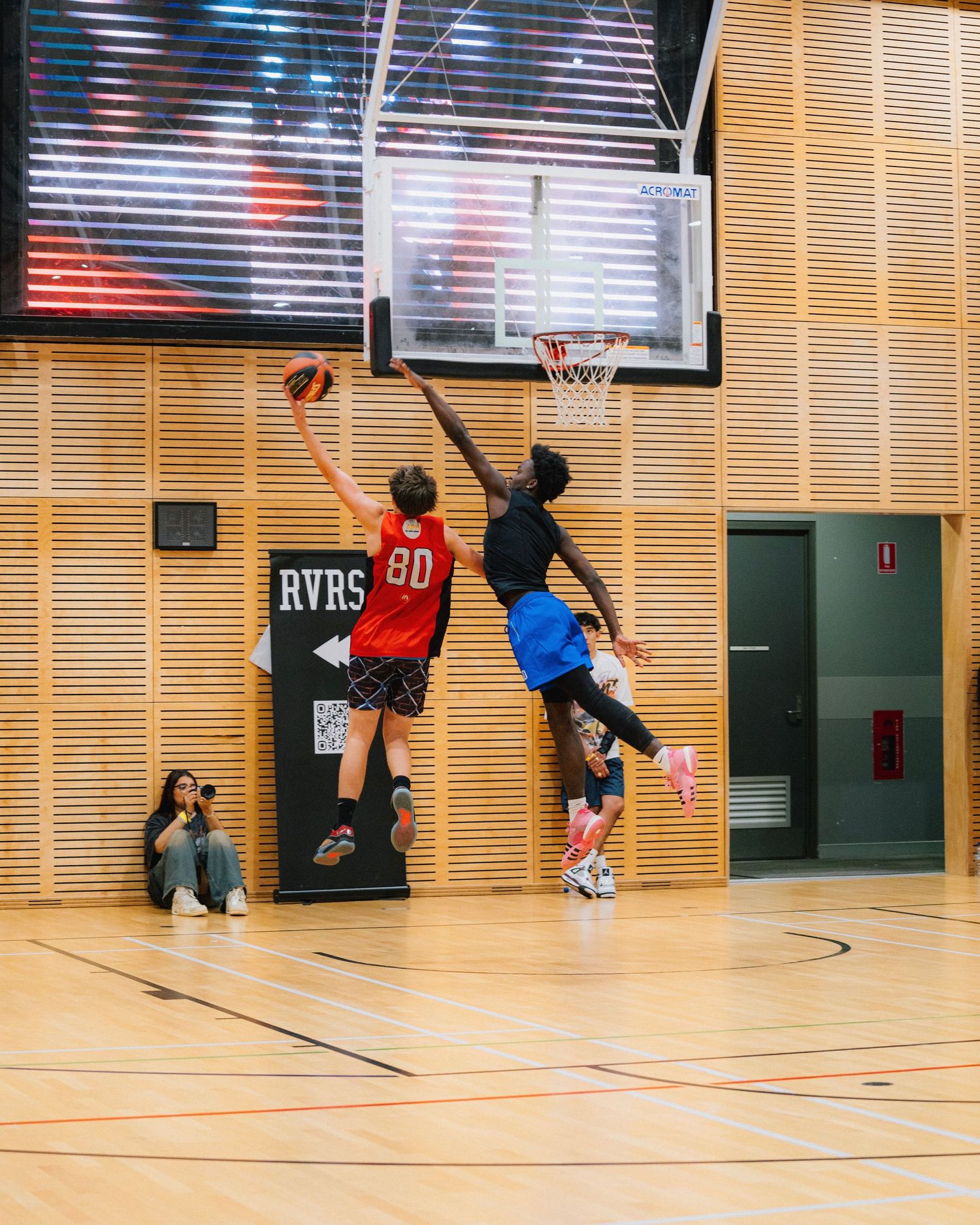 Two basketball players jumping high to compete for the ball near the hoop in an indoor gymnasium.