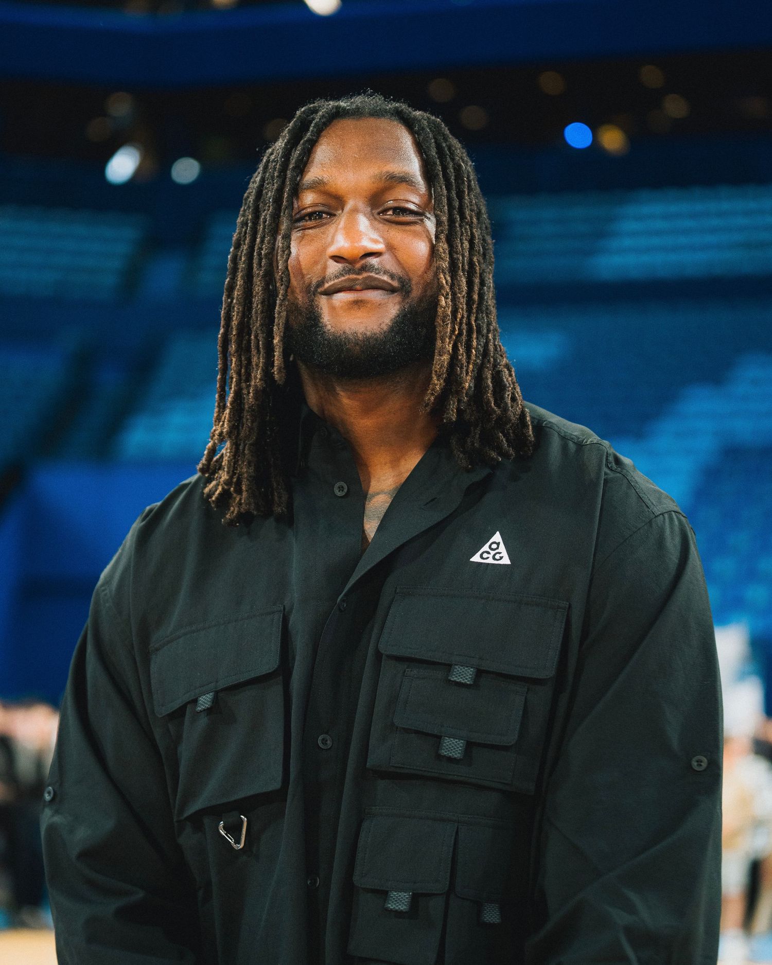 Person with dreadlocks wearing black utility jacket smiling in stadium setting.