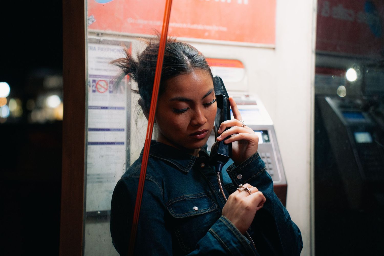 Someone in a denim jacket uses a public payphone at night in an urban setting.
