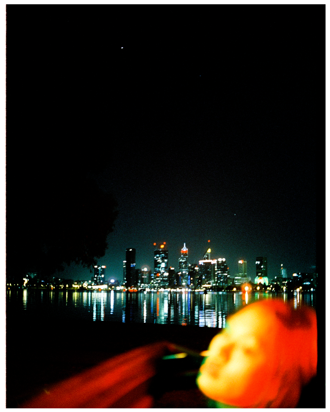 Illuminated city skyline reflects on water at night with blurred orange elements in foreground.