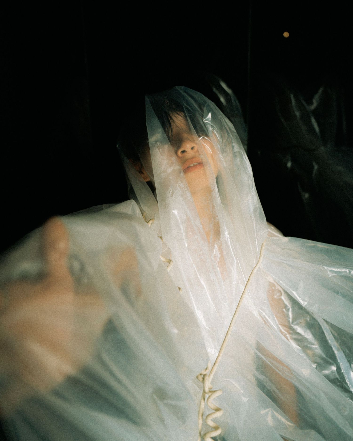 Figure draped in translucent plastic material creates ethereal artistic portrait against dark background.