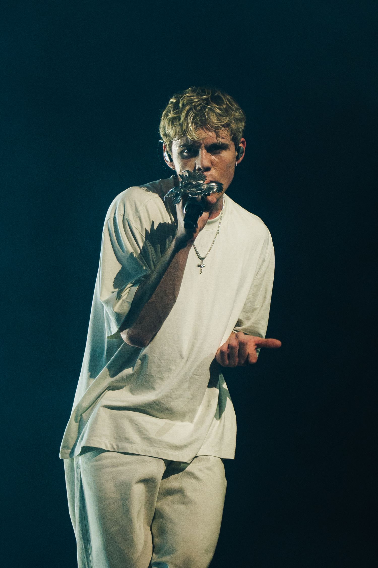 A performer in all-white clothing sings into a microphone on a darkly lit stage.