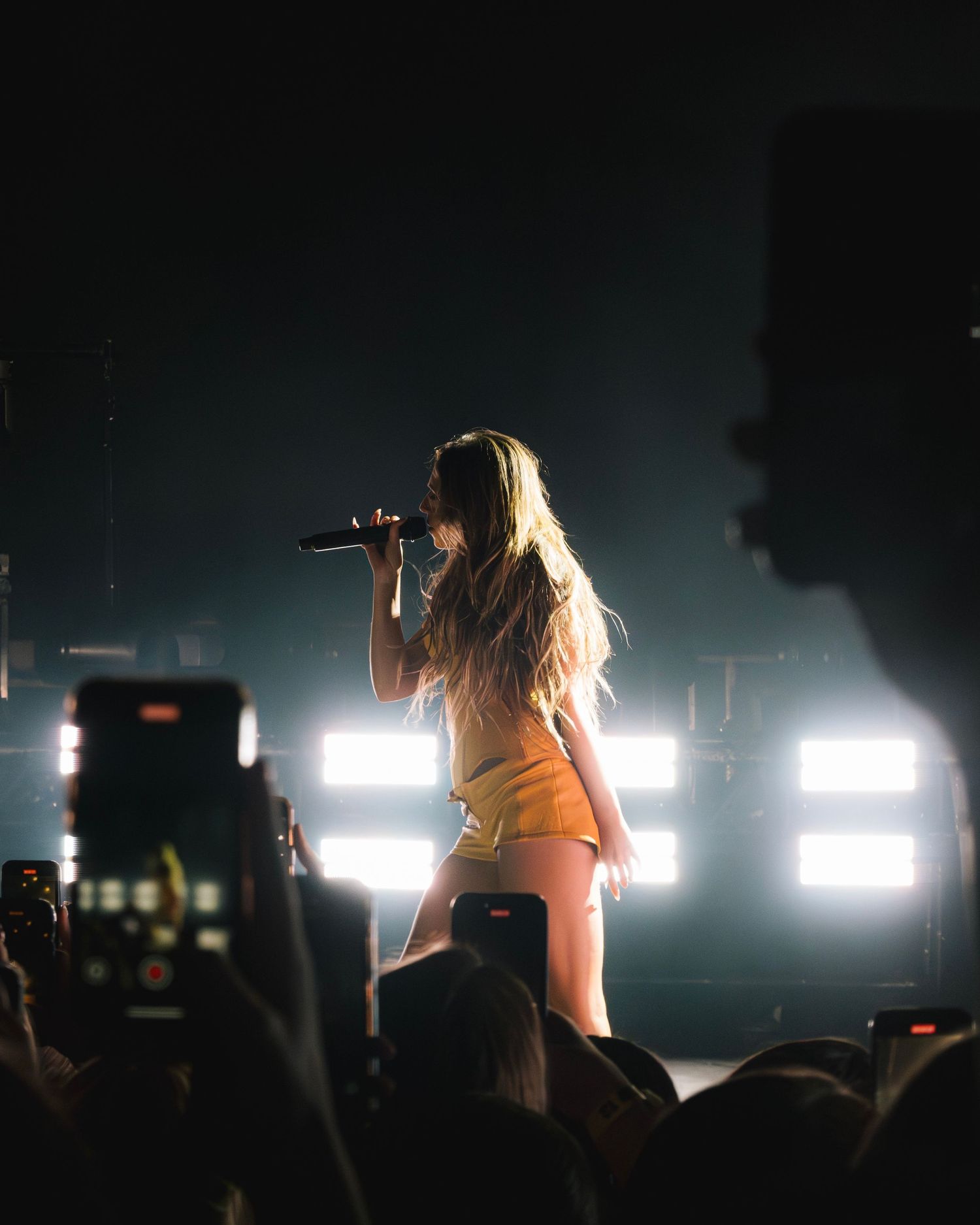 A performer on stage in white outfit sings into a microphone with bright backlights and crowd silhouettes visible.