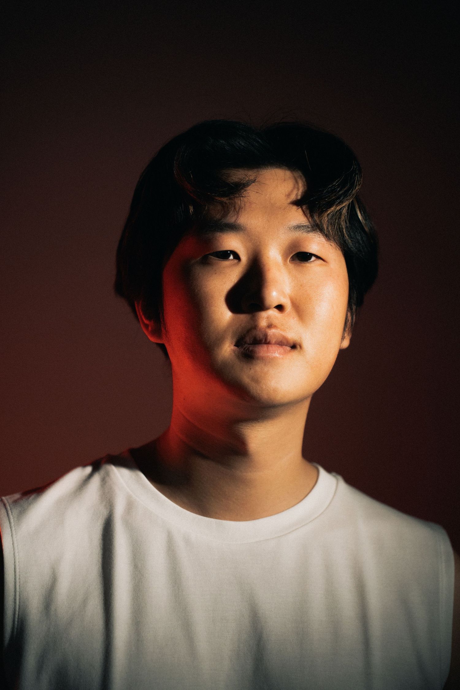 A portrait photo with red lighting on one side showing someone in a white t-shirt against a dark background.