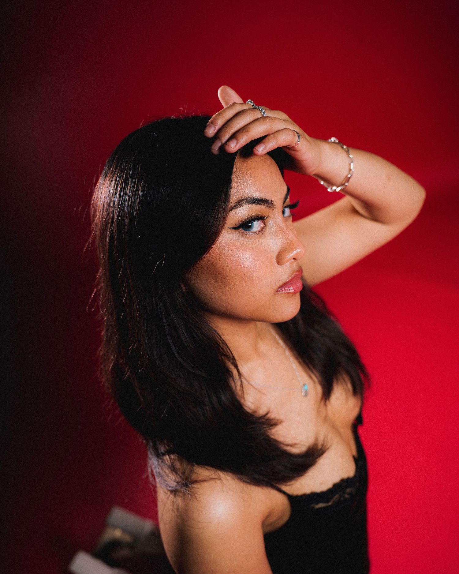 Portrait against red backdrop features long dark hair, black lace top, and delicate jewelry with dramatic pose and lighting.