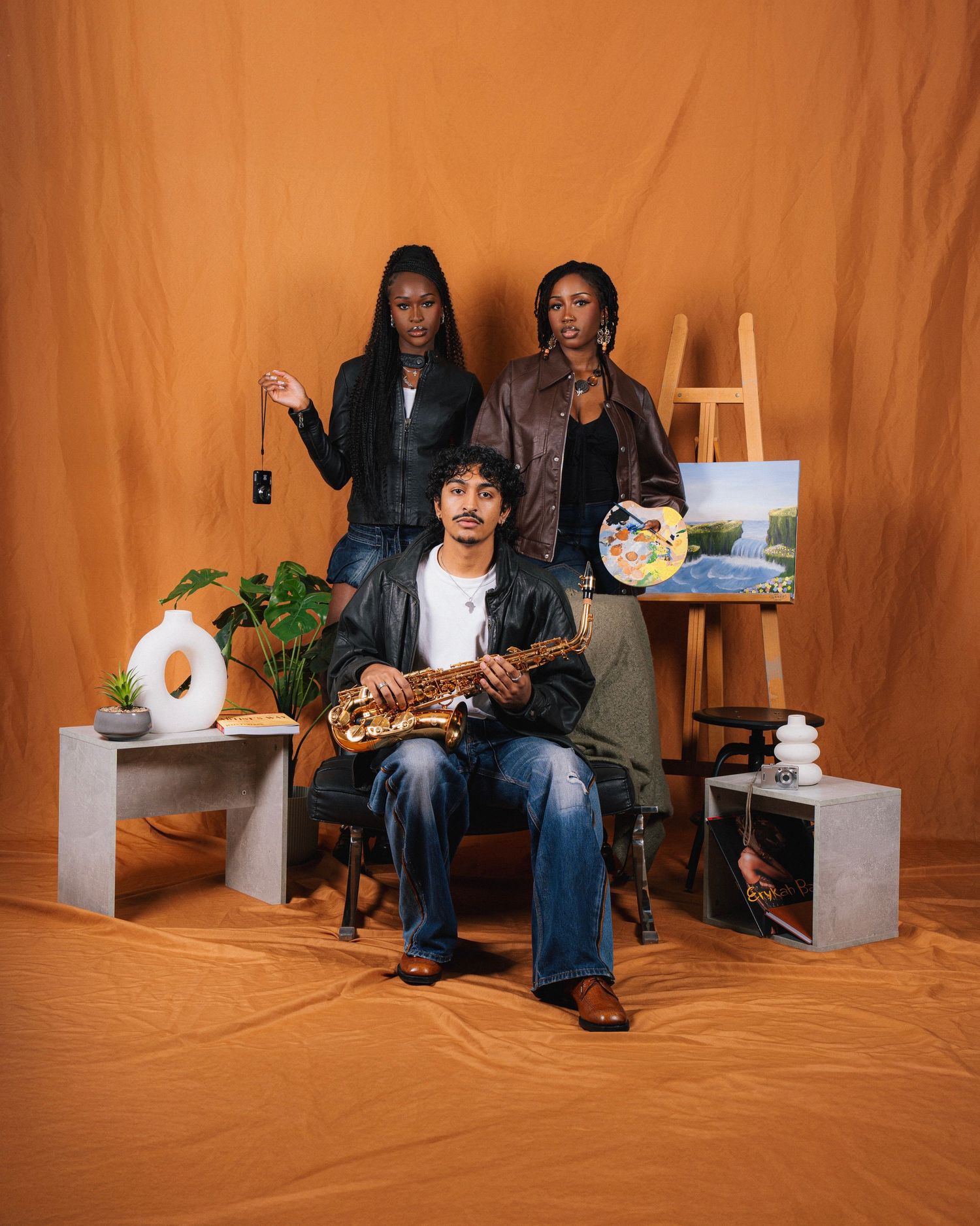 Three artists pose with creative props including a neon light, plants, and paintings against an orange backdrop in a studio setting.