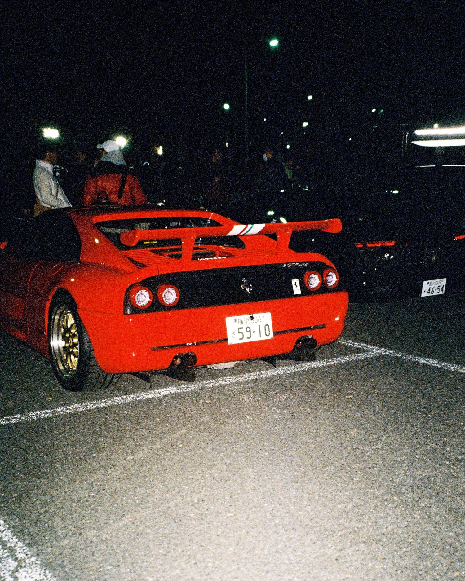 Red Ferrari F355 with aftermarket wing photographed at nighttime car meet.