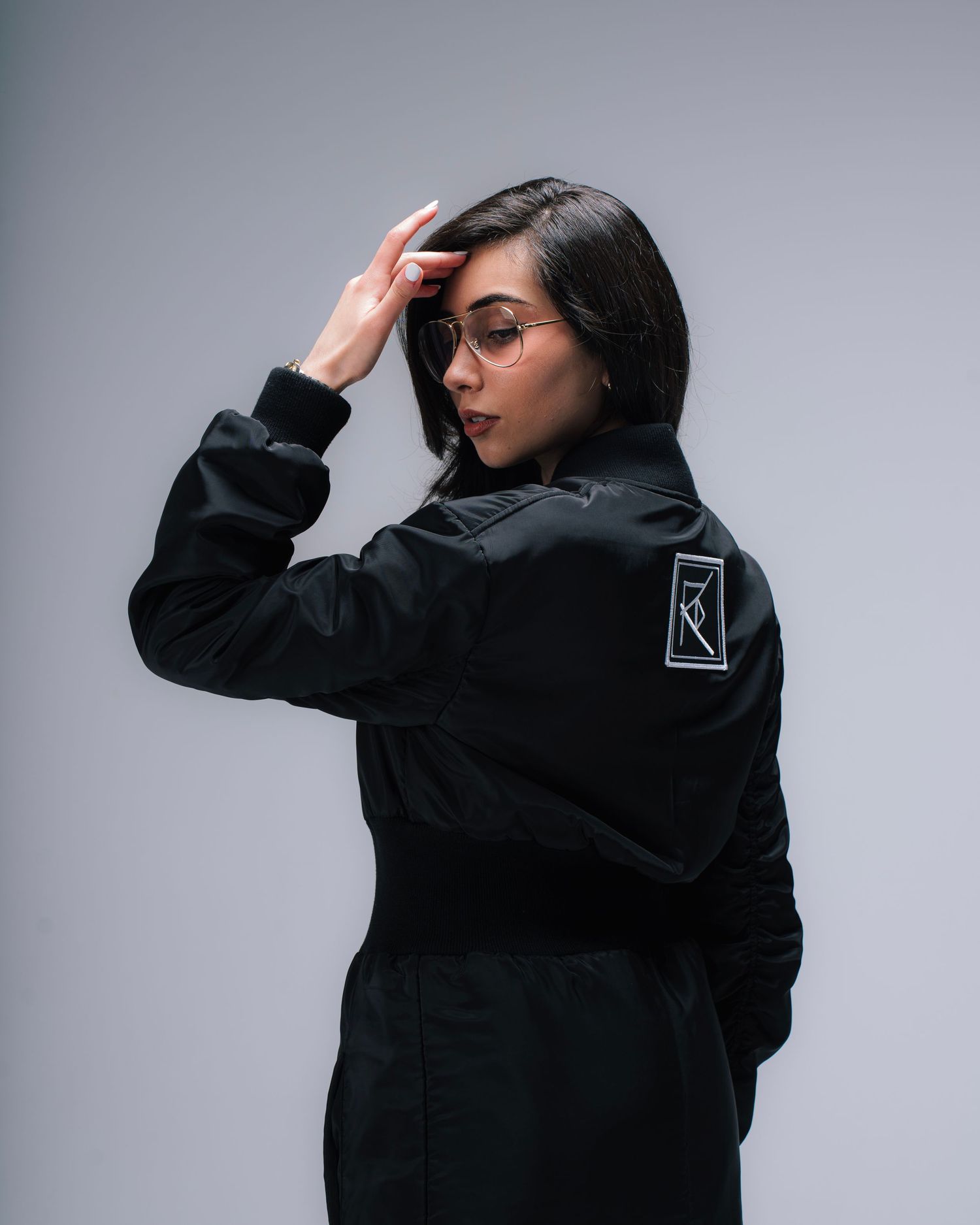 Model wearing black jacket with brand patch poses against grey background in studio.