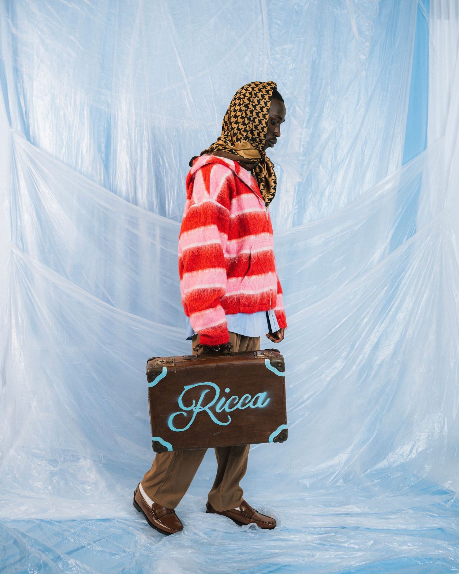Person in striped red sweater holding vintage suitcase against blue backdrop.