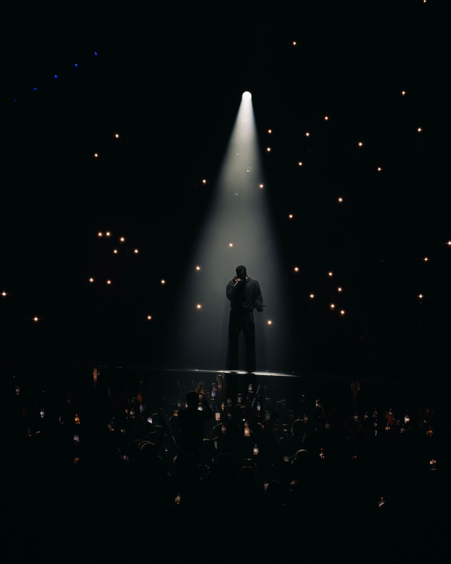 A silhouetted performer stands in a dramatic spotlight beam on stage while glowing phone lights sparkle in the dark audience.