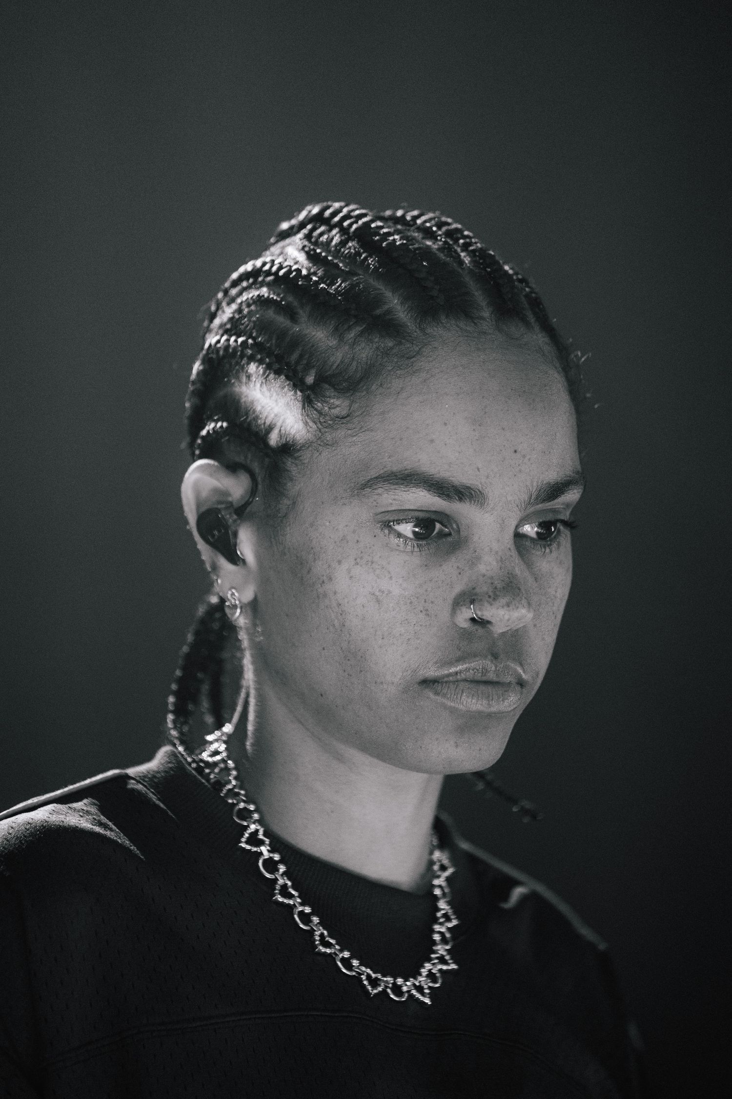 Black and white portrait showing a person with braided hair and metallic chain necklace against a dark background.