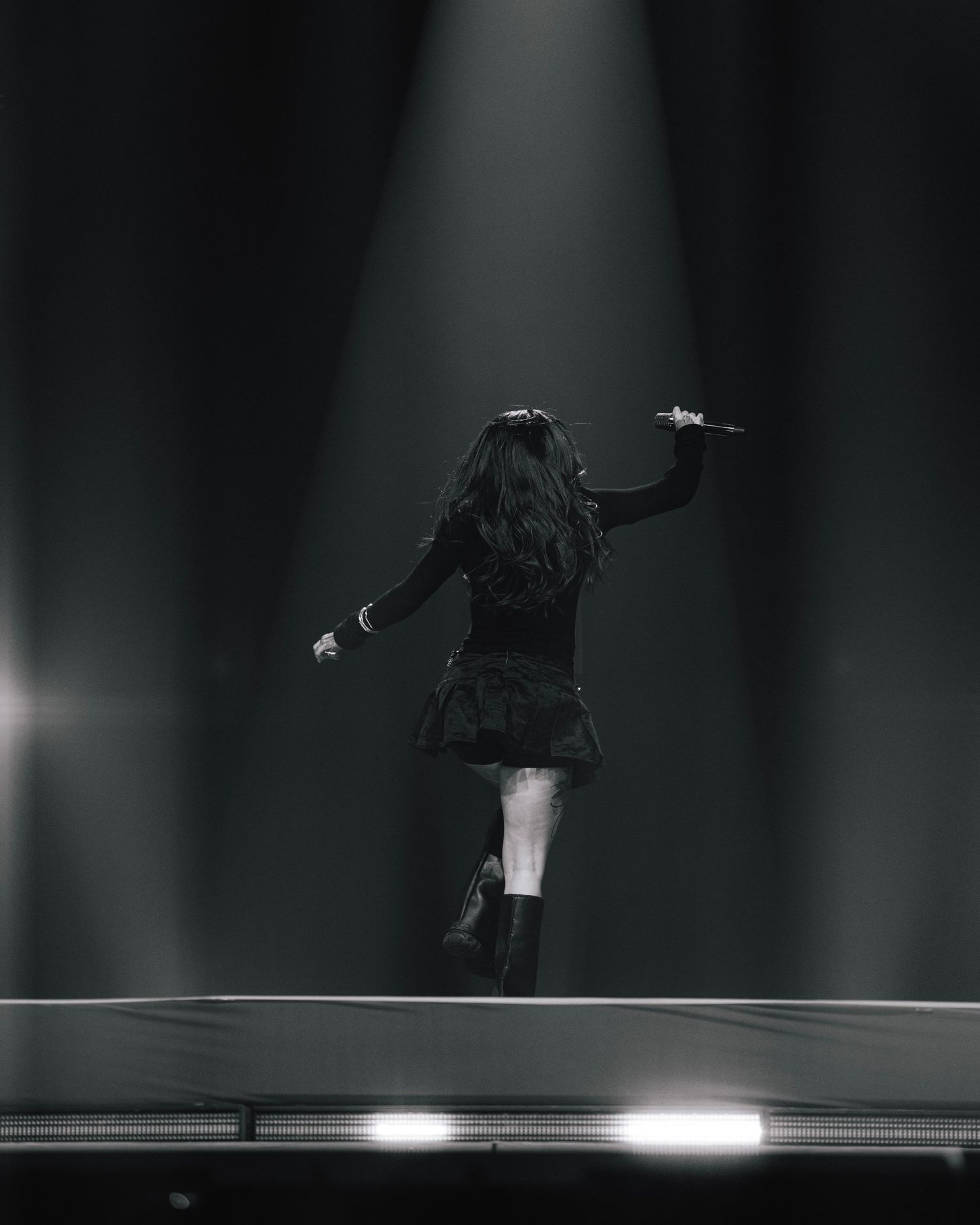 Silhouetted performer in short dress poses dynamically under dramatic spotlight on dark stage.