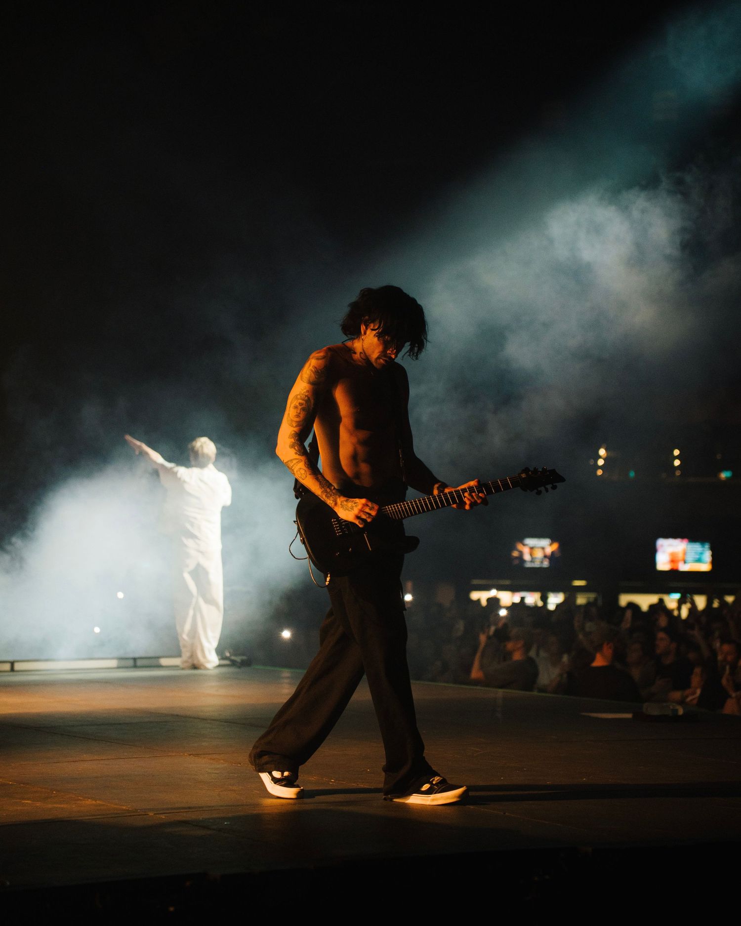 A performer in silhouette plays guitar on stage with dramatic fog and lighting effects while another figure stands in white.
