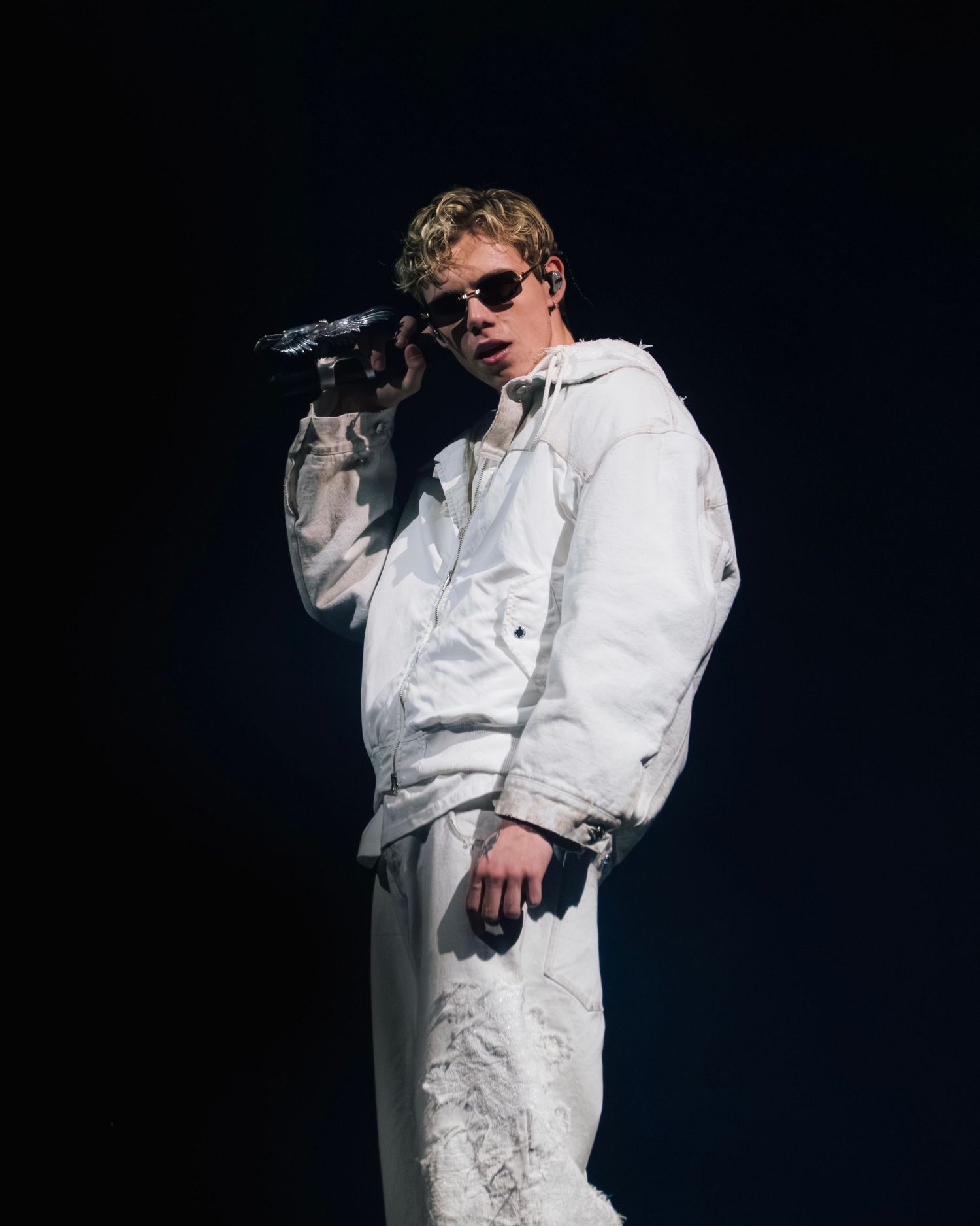 Figure in all white winter sportswear performs on stage in dramatic lighting while holding a microphone.
