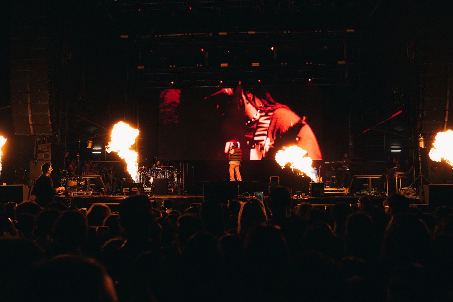 Concert stage performance with dramatic pyrotechnic flame effects and red lighting illuminating silhouettes against dark background.