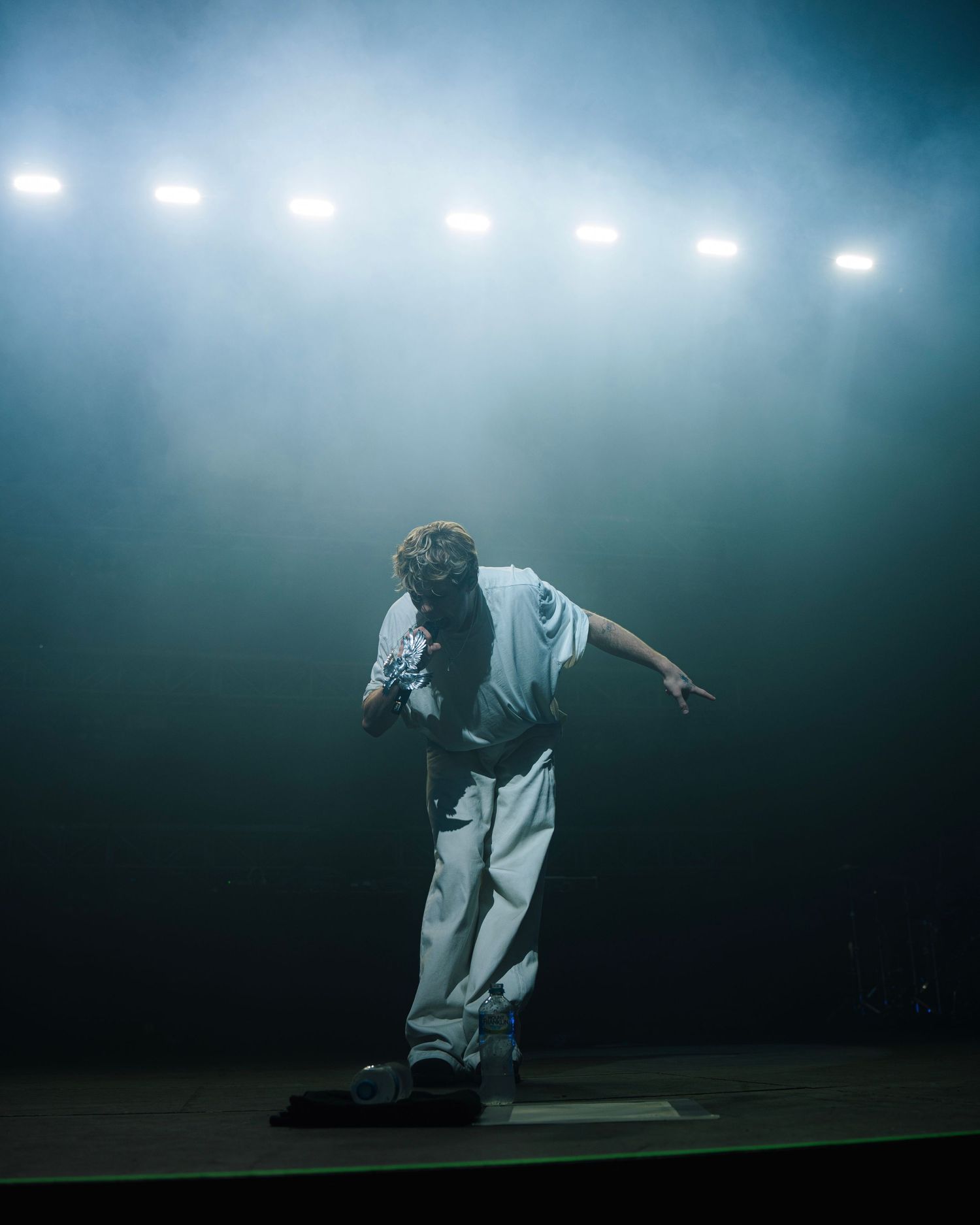 Performer in white outfit bows dramatically on dark stage with glowing stadium lights creating atmospheric fog effect above.