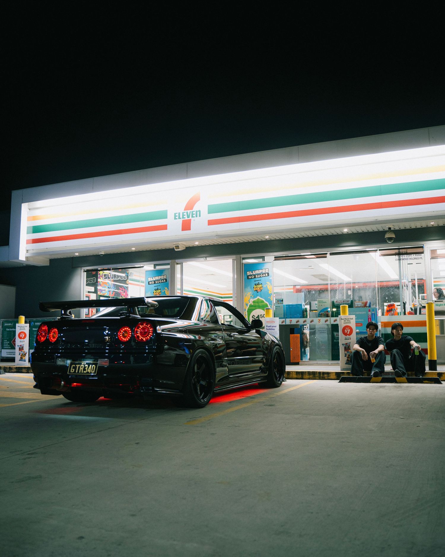 Black Nissan GTR parked at a 7-Eleven gas station at night with neon lighting.