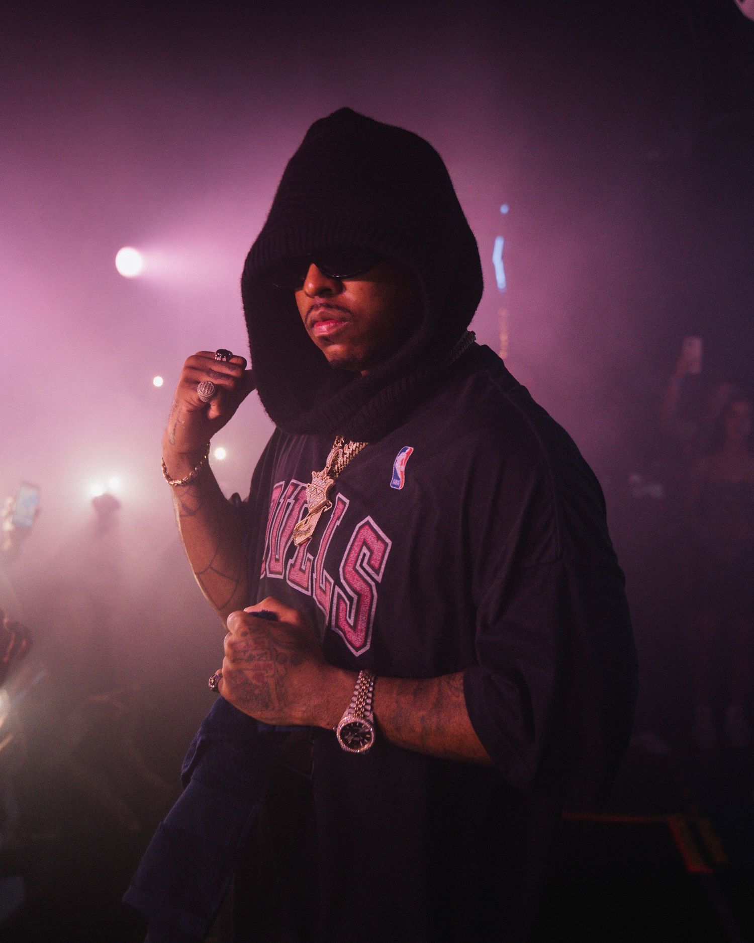Performer in hooded sweatshirt and red sunglasses performing on stage with pink lighting effects.