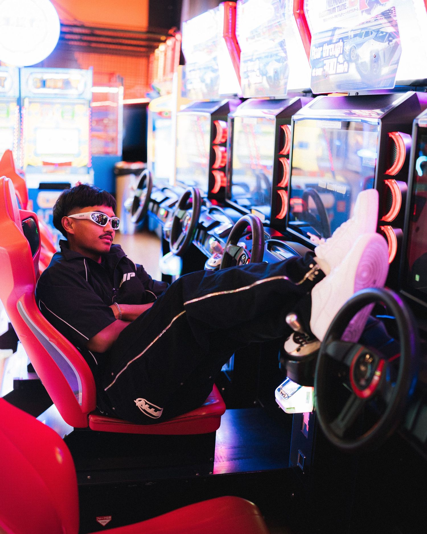 Young person in sunglasses enjoying virtual racing game in arcade with glowing neon lights and racing seat simulator.