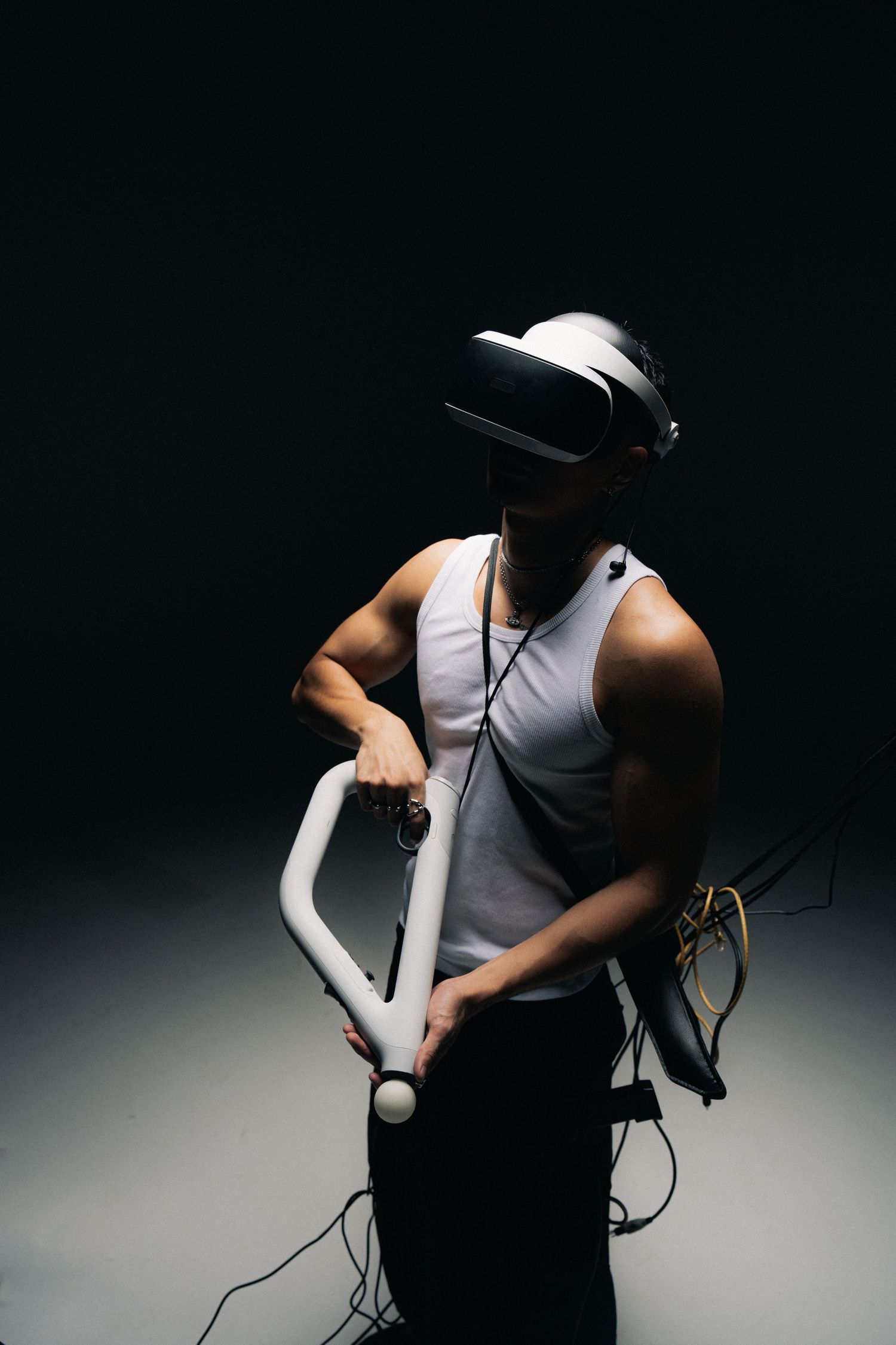 Person wearing VR headset and white tank top holds gaming controller against dark background in dramatic lighting.