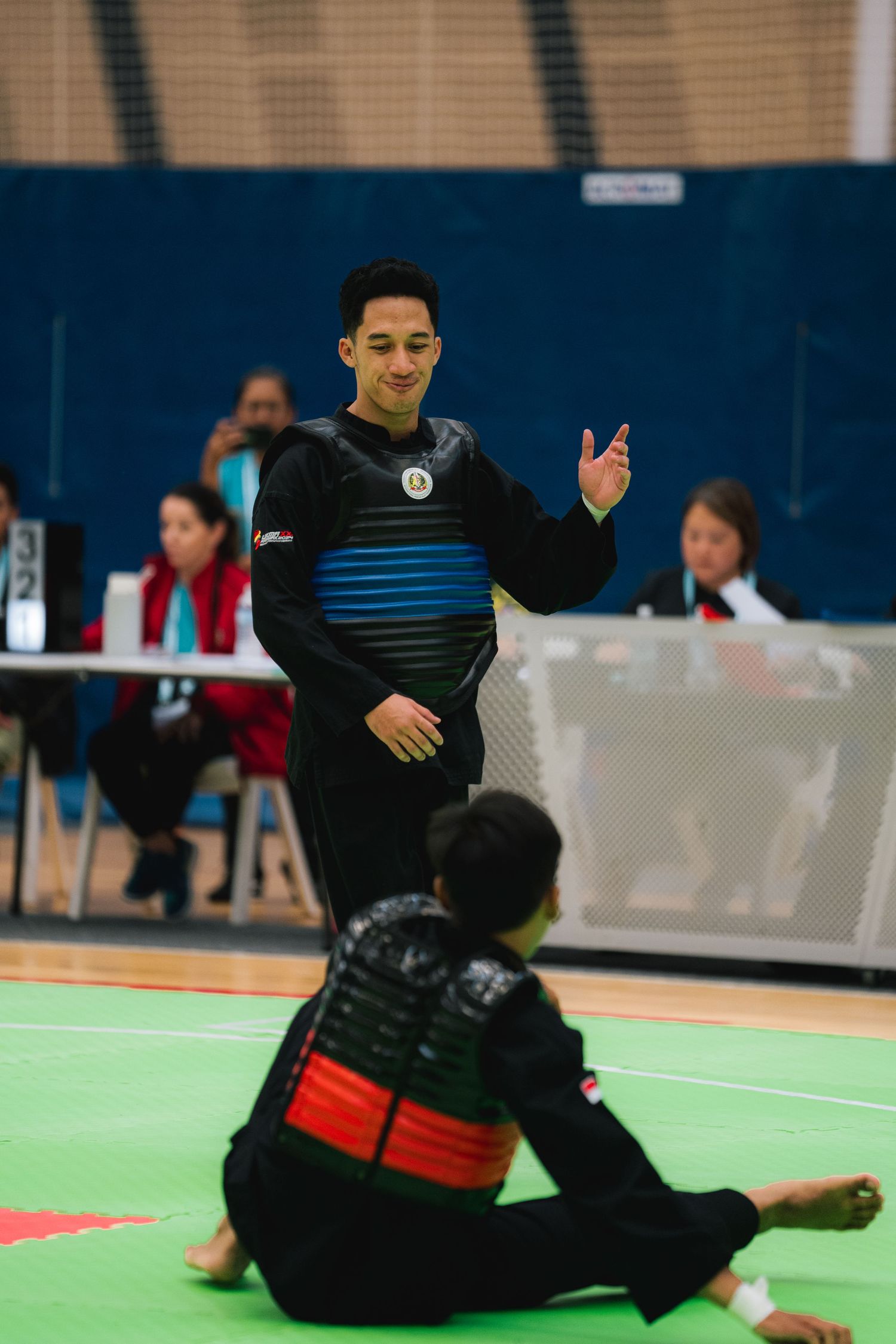 Martial artists perform in a competition on a green mat floor with judges watching in the background.